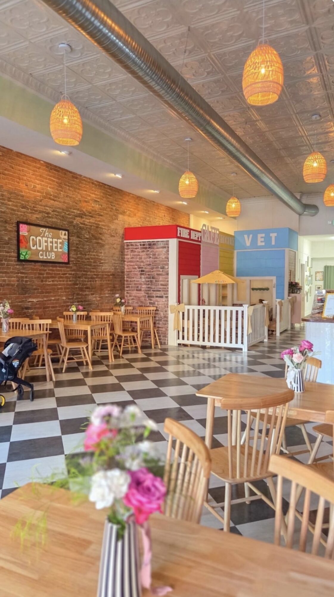 Café interior with wooden tables and chairs, hanging orange lights, checkered floor, brick wall, and a pet vet area in the background.