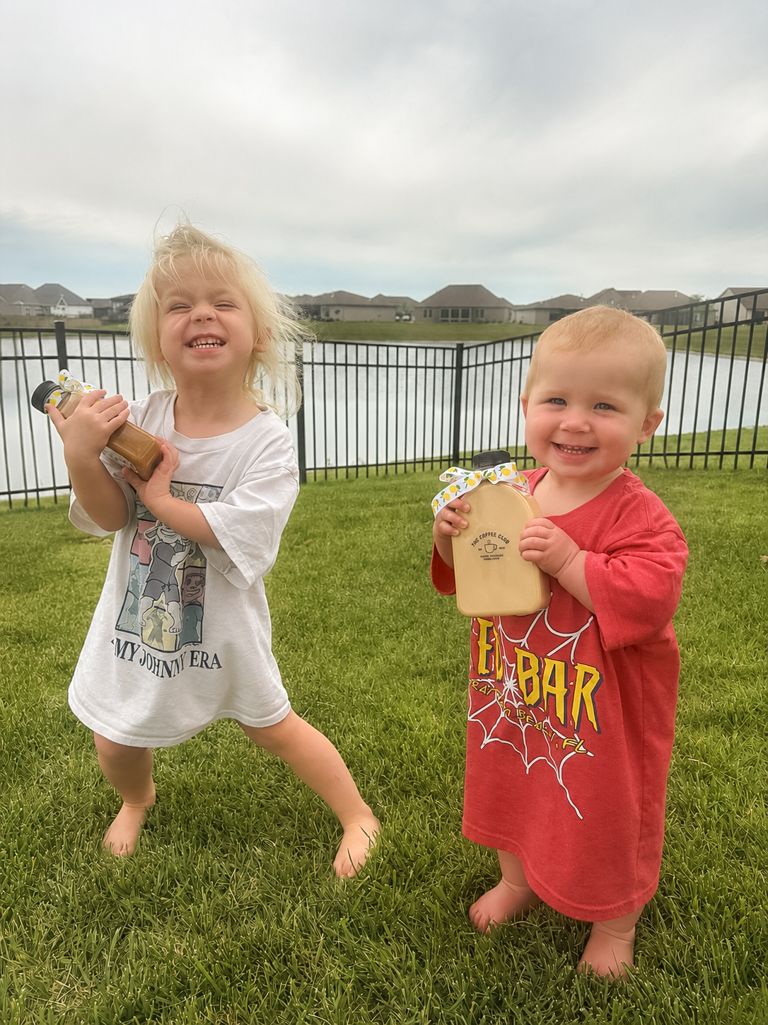 Two children standing on grass near a fence, holding drinks, smiling, with a lake and houses in the background.