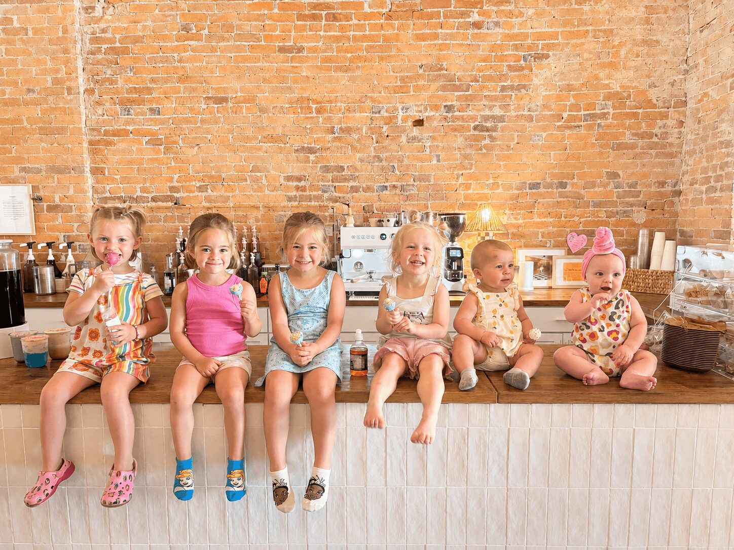 Six children sitting on a wooden counter in front of a brick wall, some holding drinks, with a kitchen background.