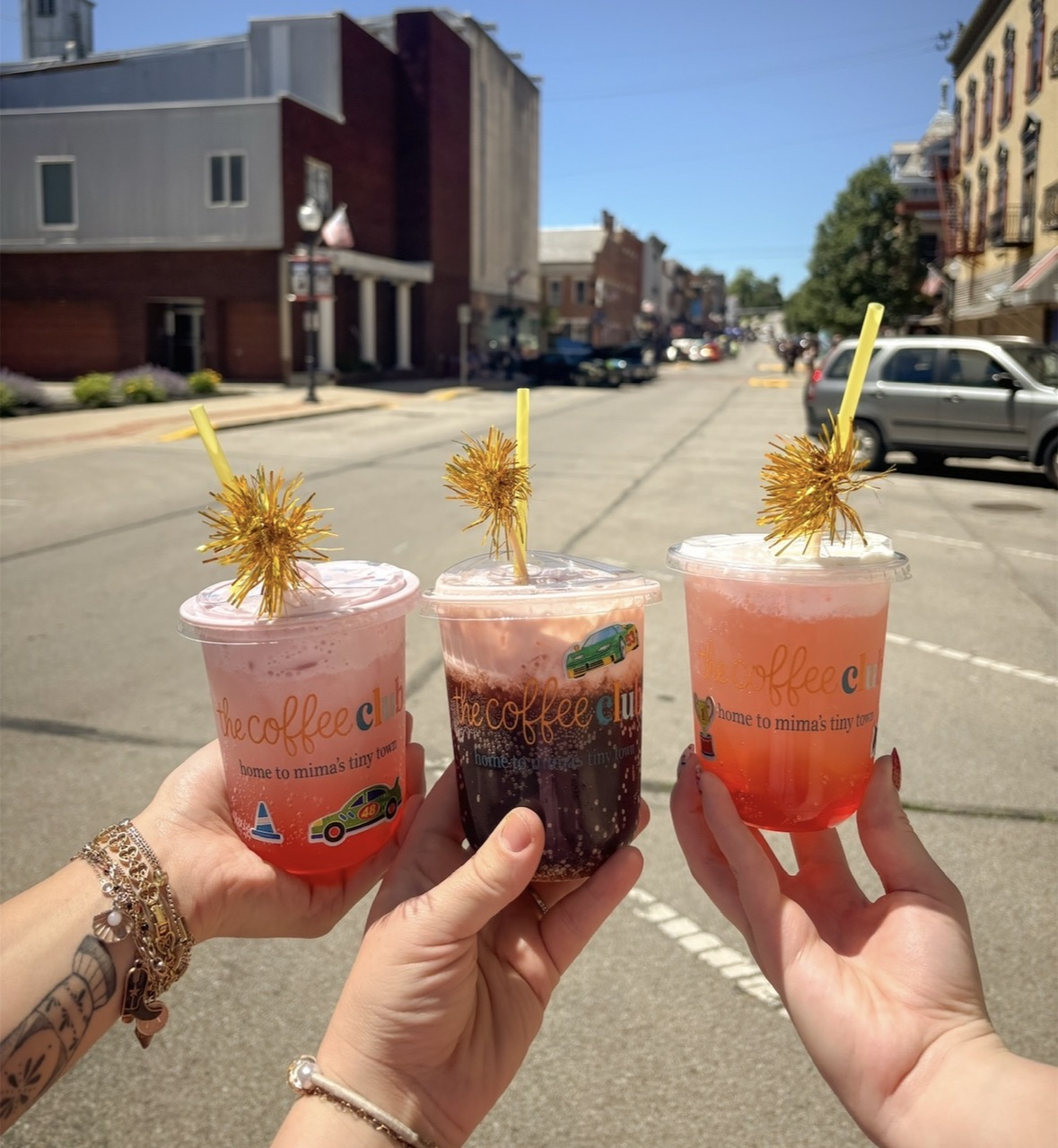 Three people hold colorful drinks with yellow straws and decorations on a city street, buildings and cars in background.