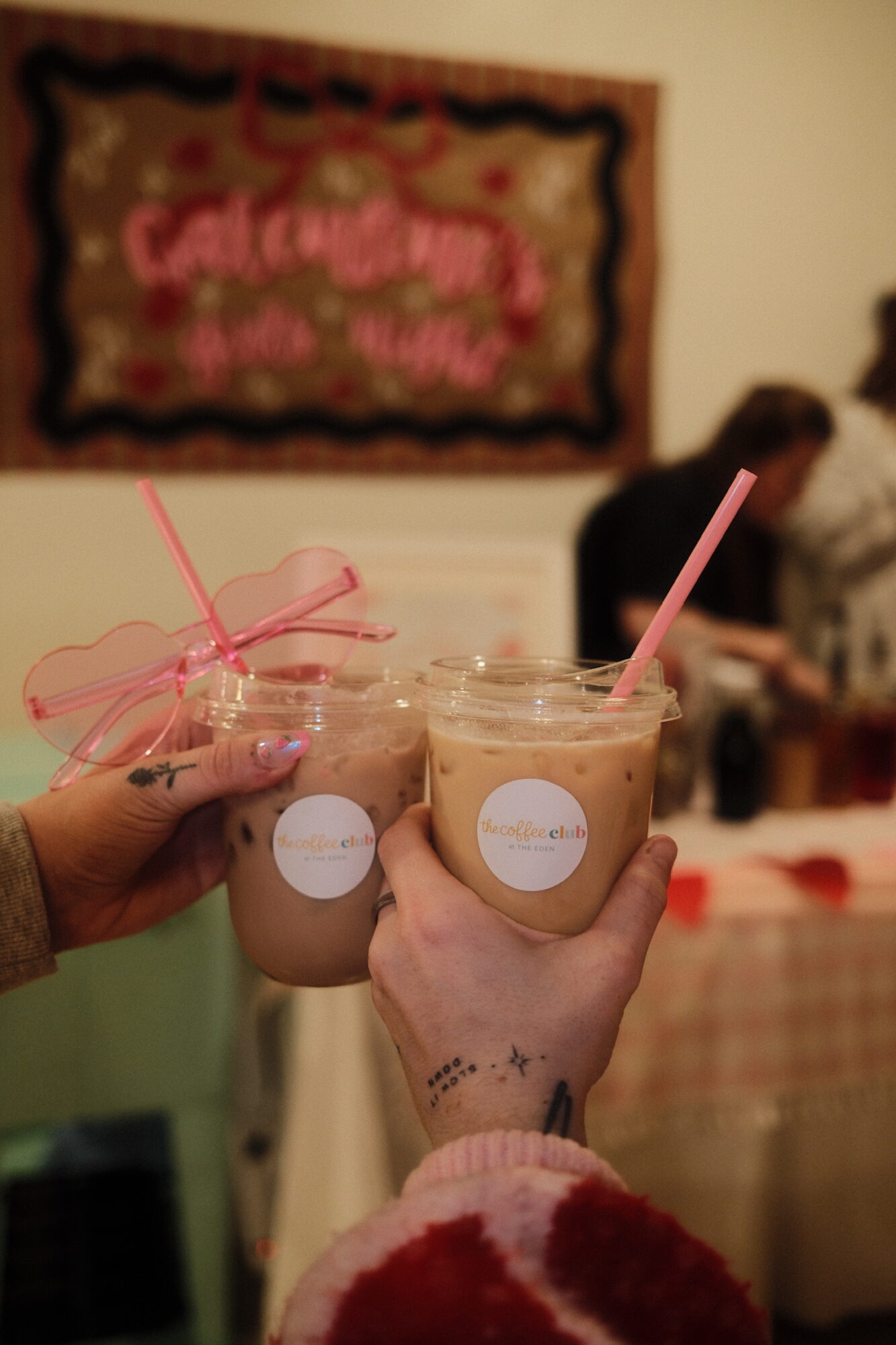 Two hands holding glasses of iced coffee with pink straws, background shows a blurred person and a decorative wall hanging.