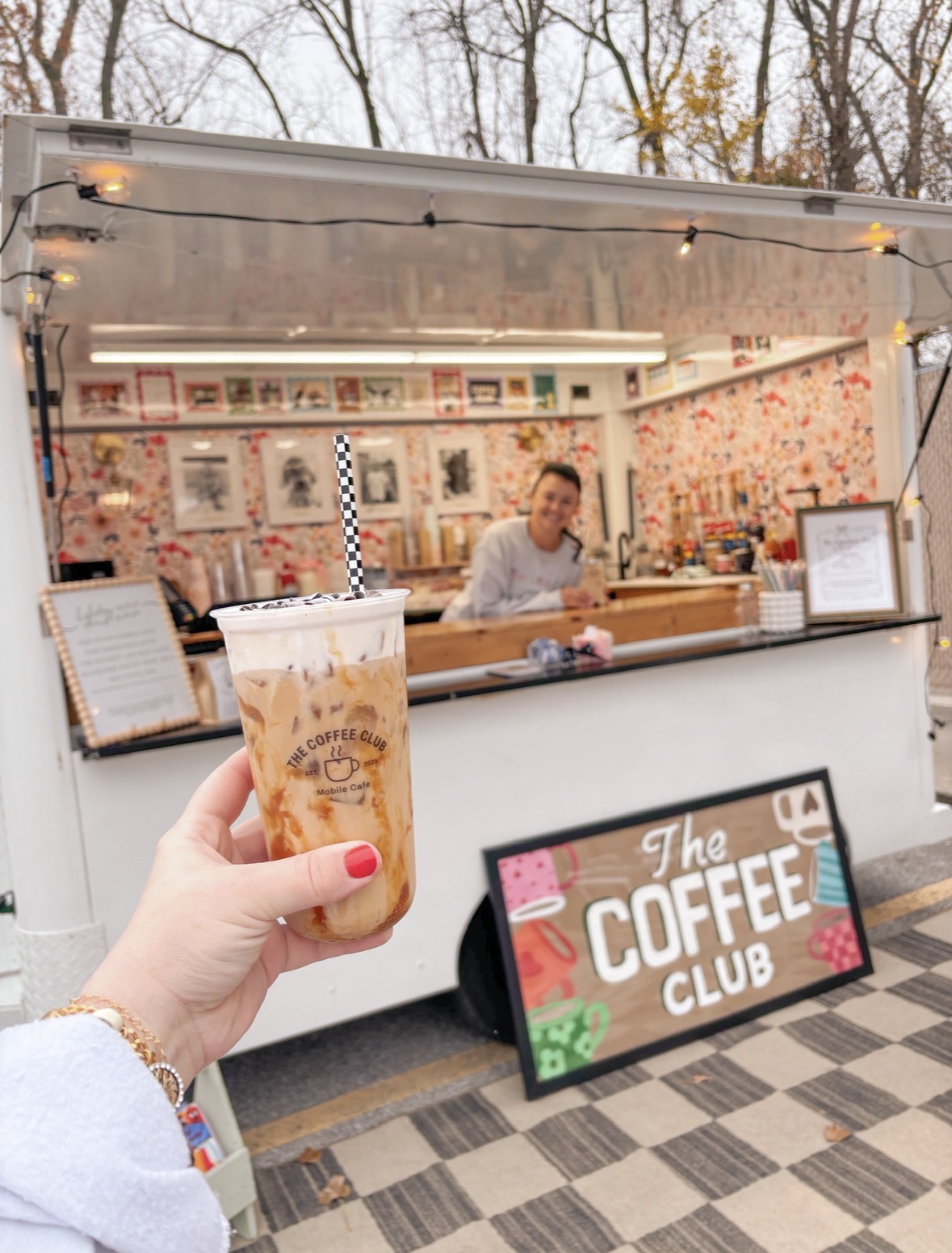 Person holding iced coffee with a barista in a coffee stand behind the counter outdoors.