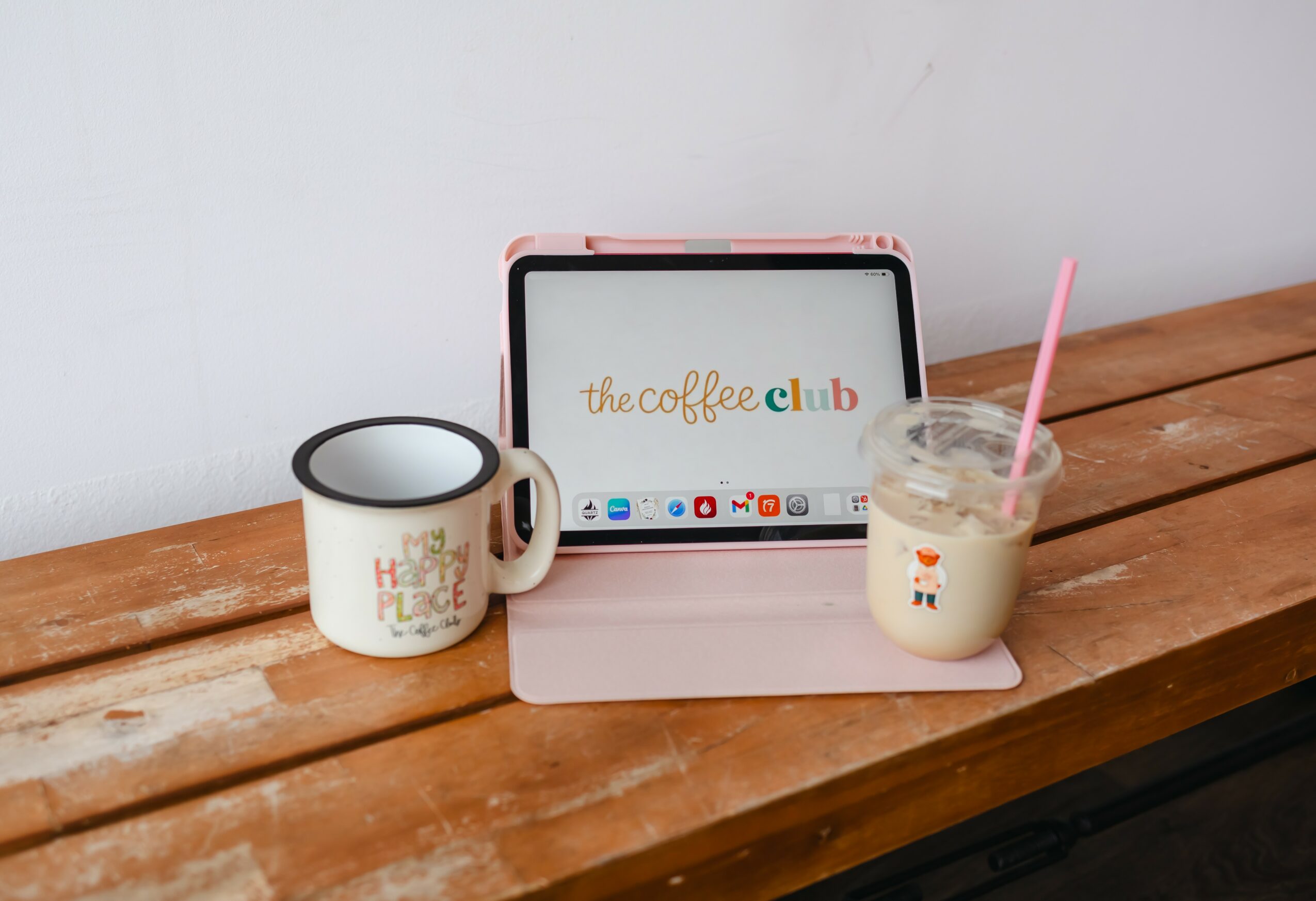 Tablet on pink stand displaying 'the coffee club' logo, mug with text, and a glass of iced coffee with a pink straw on wooden surface.