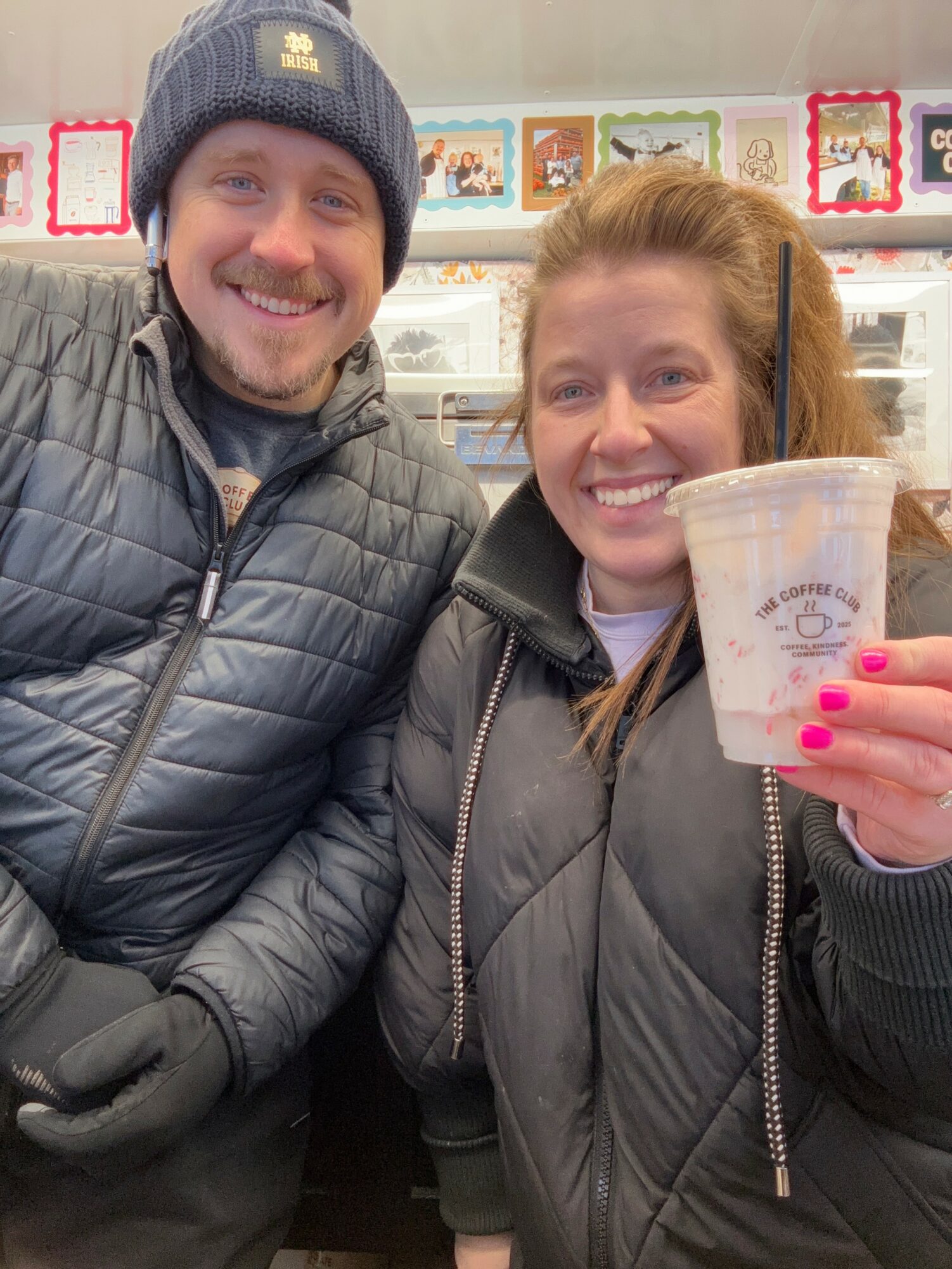 Two smiling people in winter jackets, one holding a drink, standing in a store with greeting cards in the background.