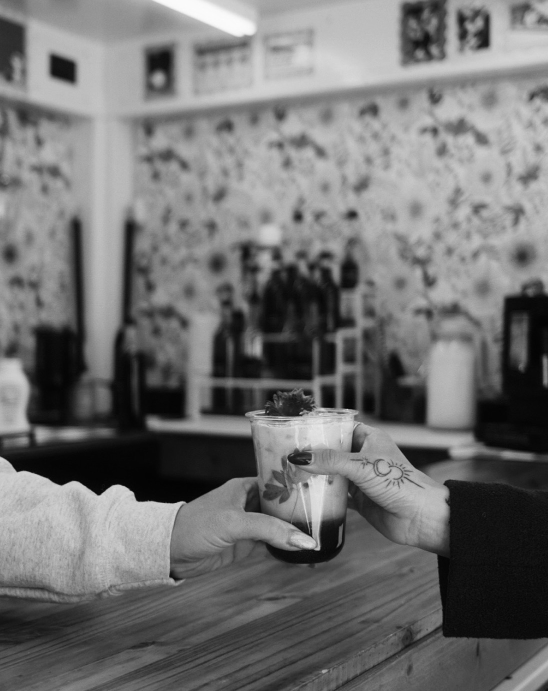 Two hands exchanging a glass of layered drink in a room with patterned wallpaper and shelves.