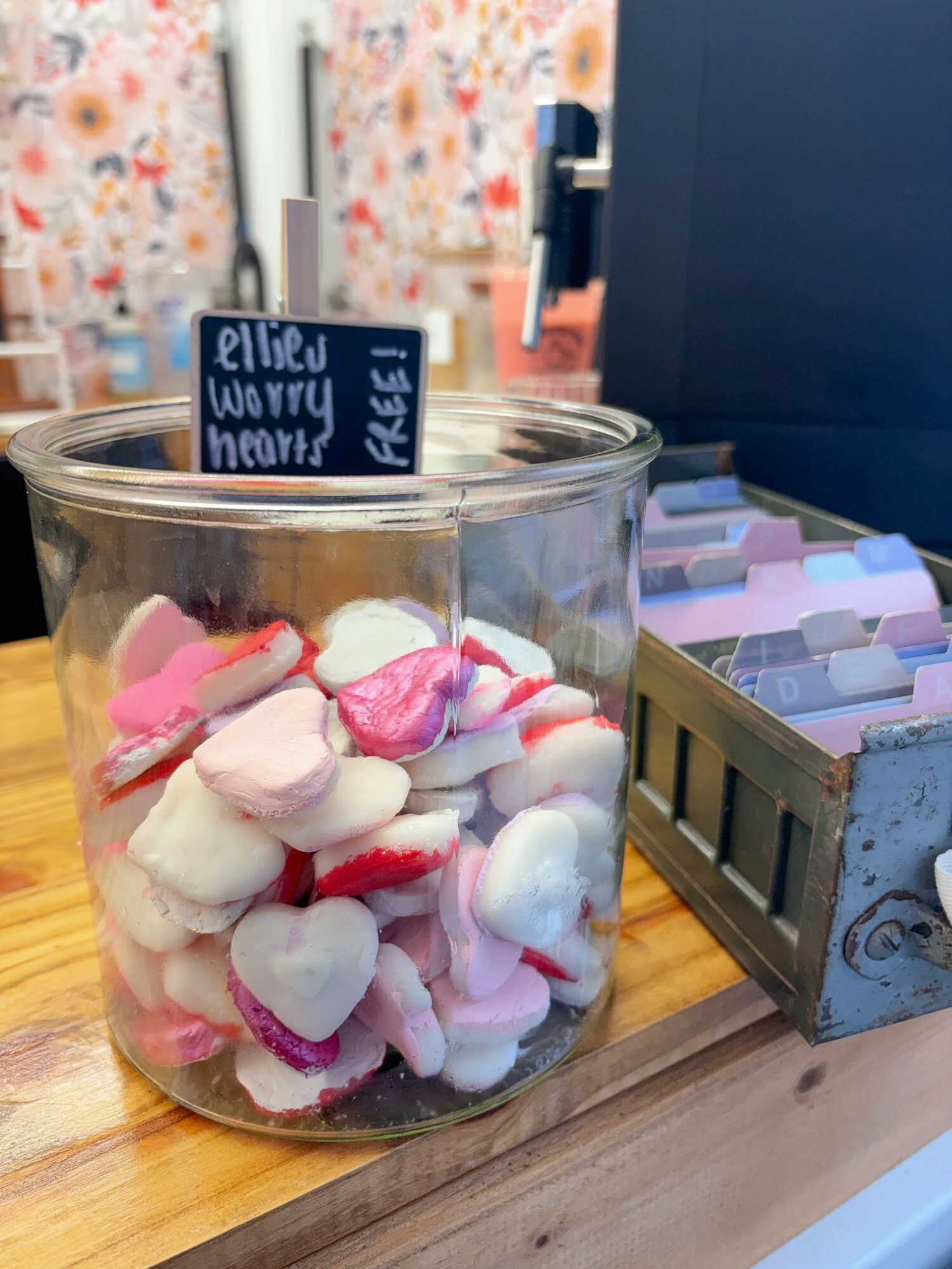 Glass jar filled with pink and white heart-shaped candies on a wooden surface, with a box of pastel-colored items nearby.