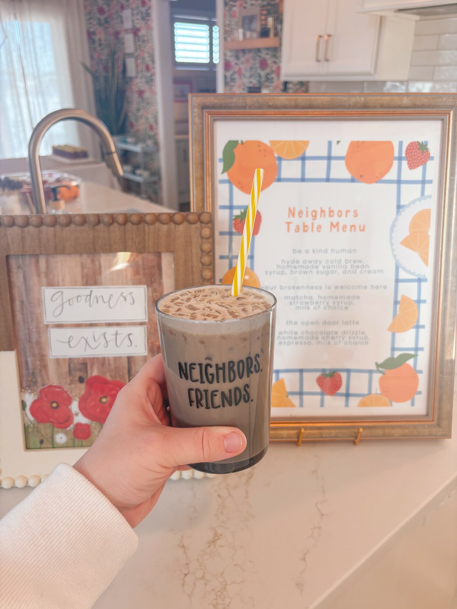 Hand holding a coffee cup with a yellow straw, labeled 'Neighbors & Friends,' in front of a framed menu and a bag on a counter.