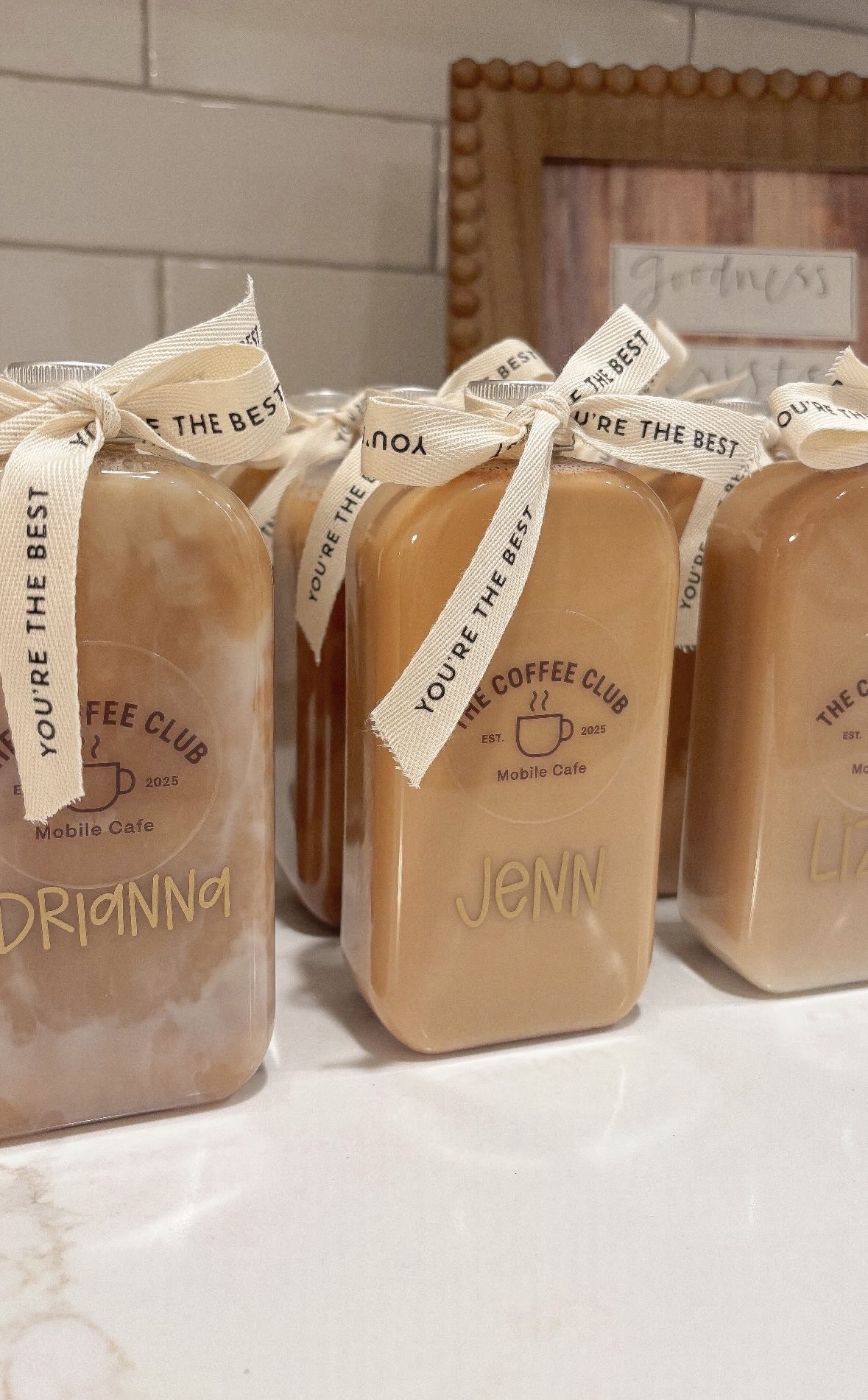 Three bottles of coffee syrup with ribbons and labels, placed on a white surface against a brick wall background.