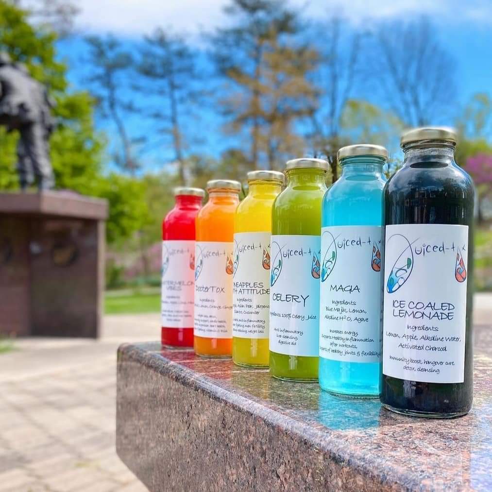 Six colorful bottles of juice or lemonade lined up on a stone surface outdoors with trees and blue sky in background.