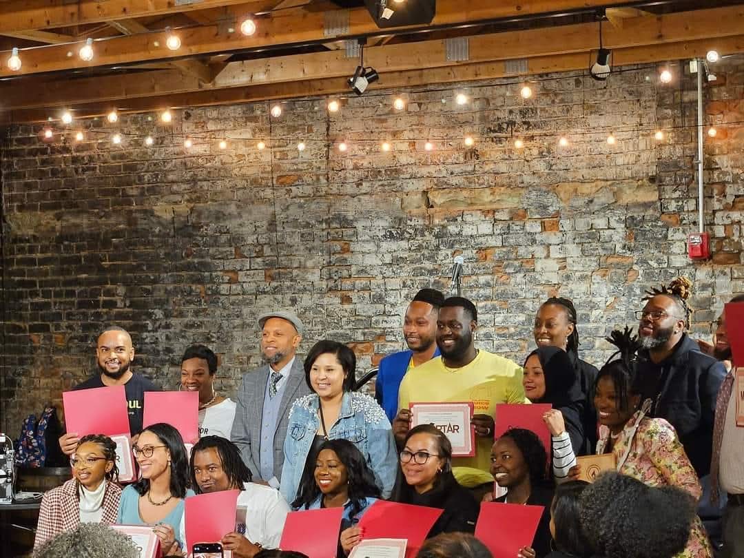 Group of people standing and sitting in a room with a brick wall and string lights, holding pink and red papers.