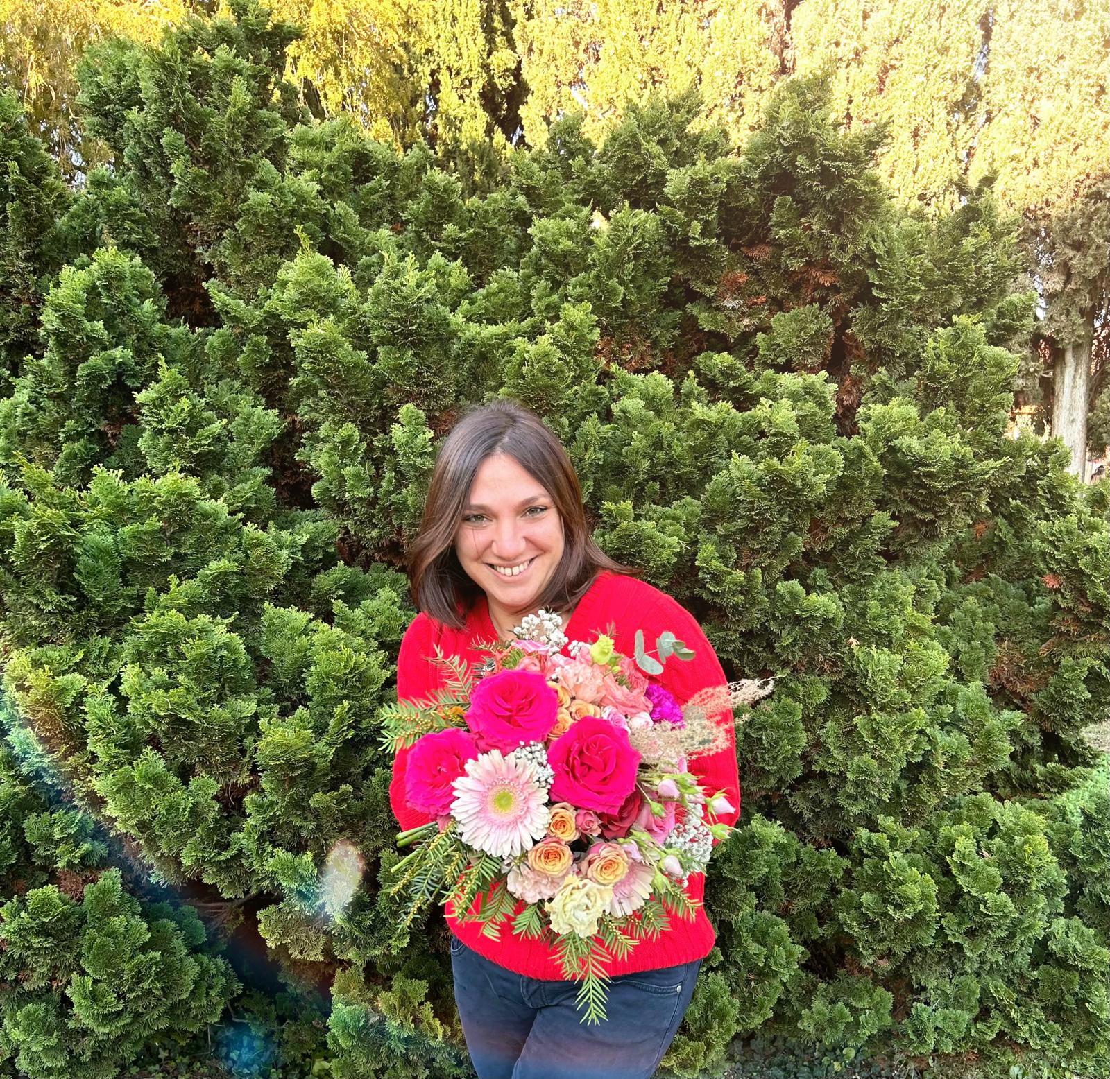 Smiling woman holding a large colorful flower bouquet outdoors, surrounded by green bushes.