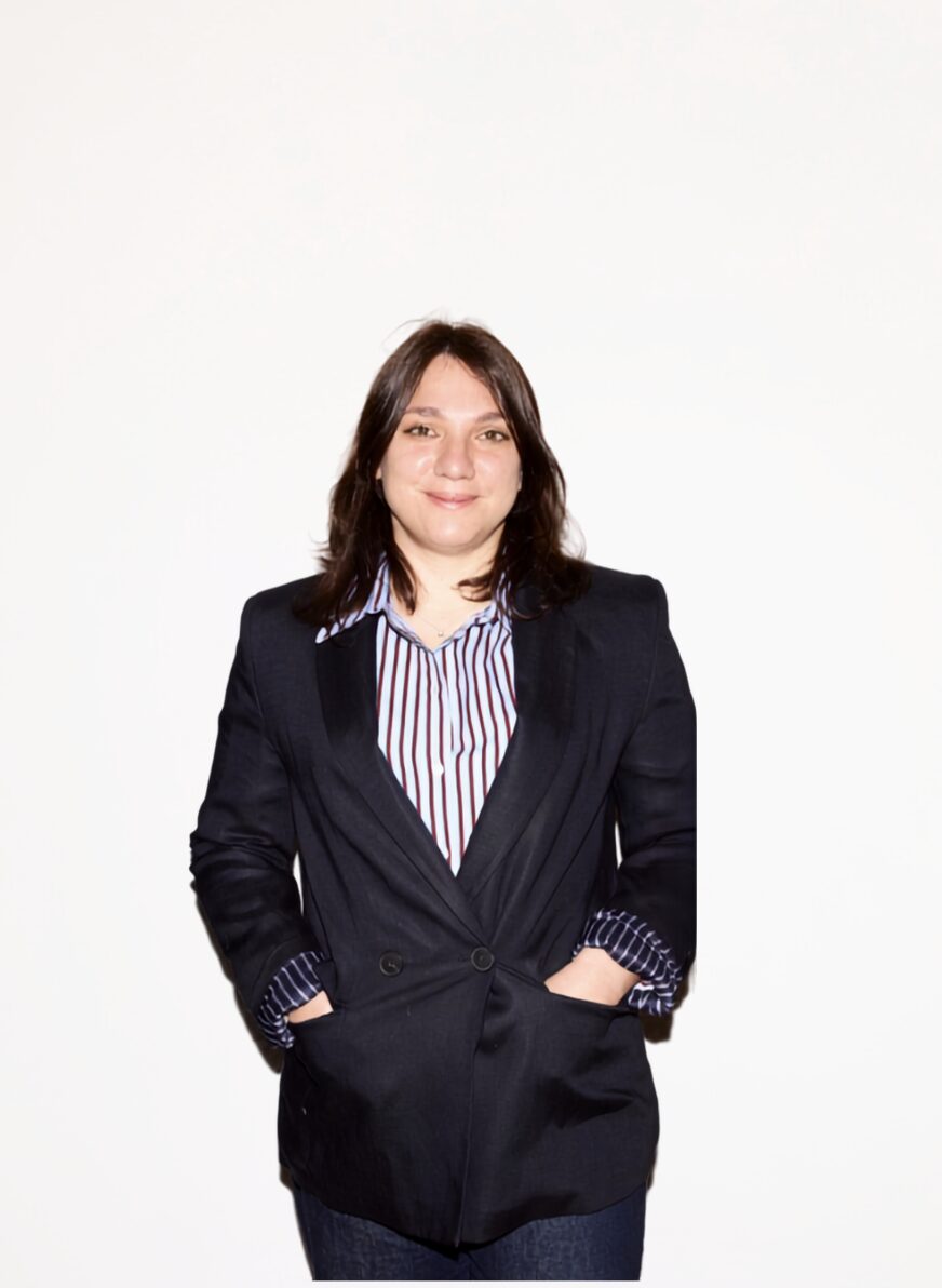 Woman with shoulder-length dark hair wearing a dark blazer and striped shirt, standing with hands in pockets against a white background.