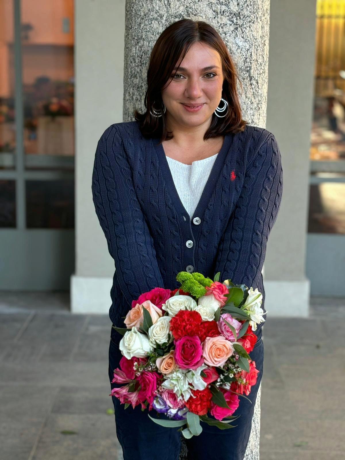 Young woman with shoulder-length dark hair holding a colorful bouquet of flowers, standing outdoors near a tree.