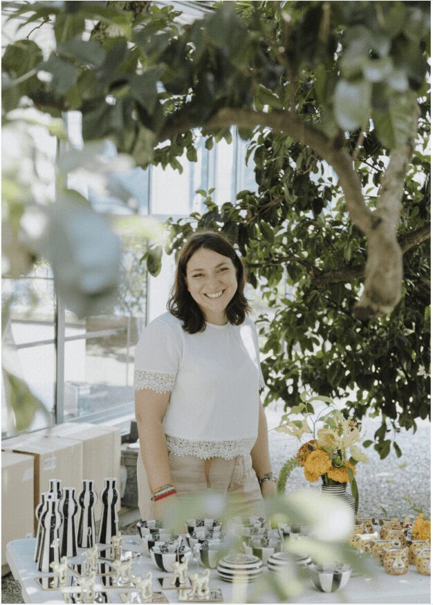 Smiling woman standing indoors near a table with flowers and dishes, surrounded by leafy trees and natural light.