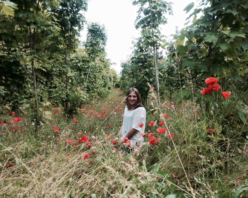 Woman standing among tall grass and red flowers in a green, wooded area.