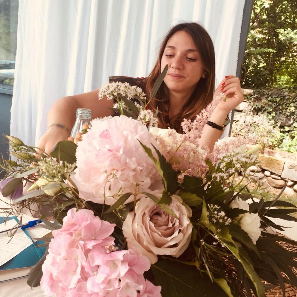 Young woman with long brown hair sitting behind a large bouquet of pink and white flowers, smiling gently, with a window and greenery in the background.
