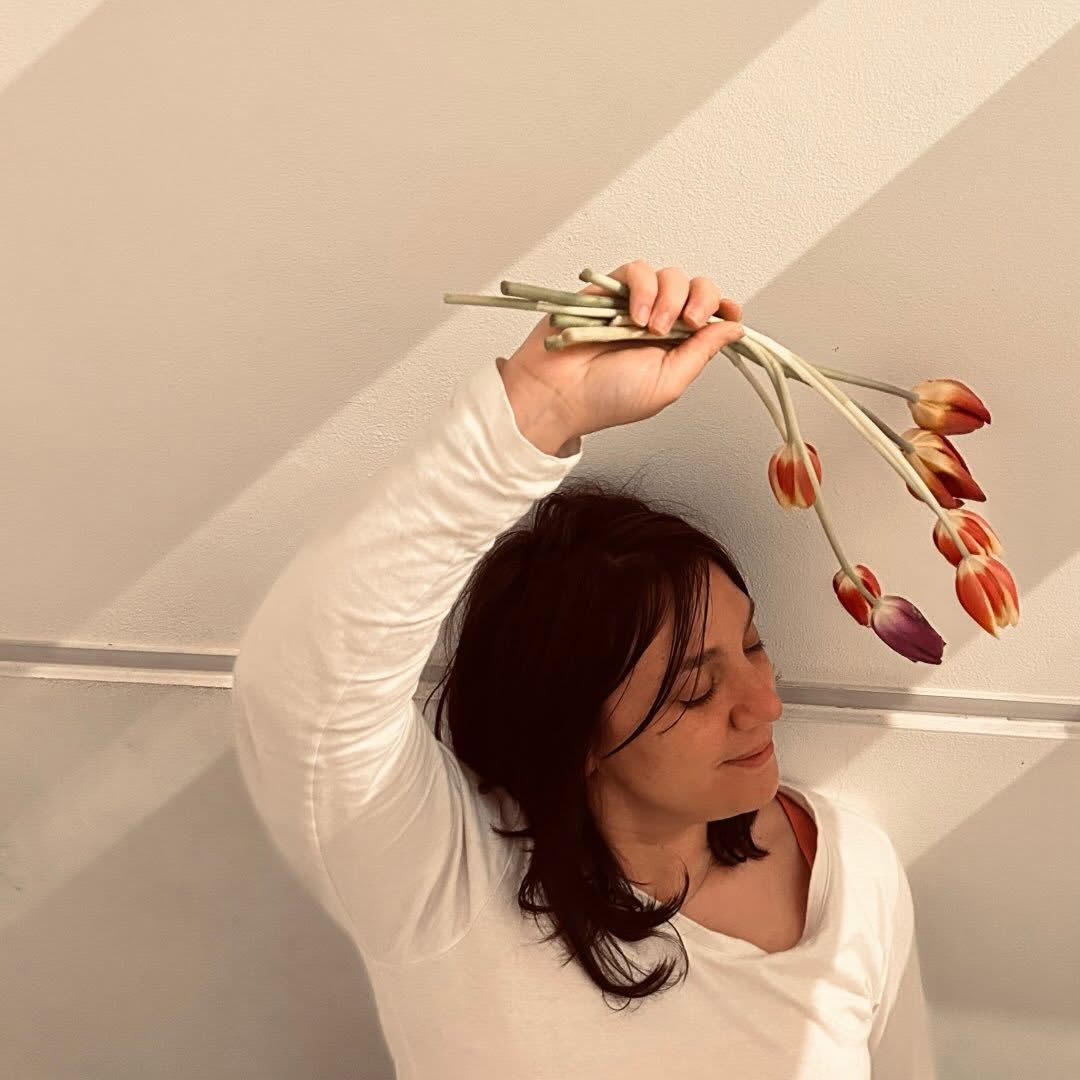 Woman with dark hair in a white shirt holds a bouquet of tulips above her head, against a white wall.