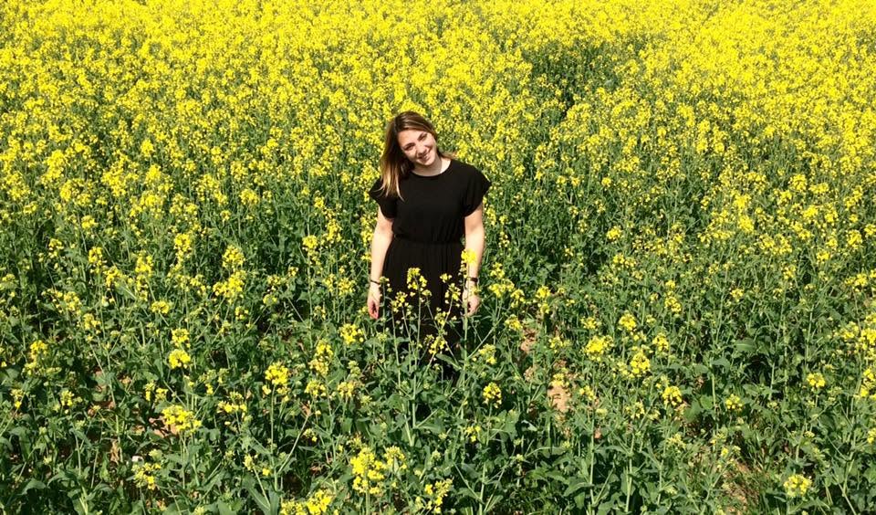 Woman standing in a yellow flower field, smiling, wearing a black dress, surrounded by blooming plants.
