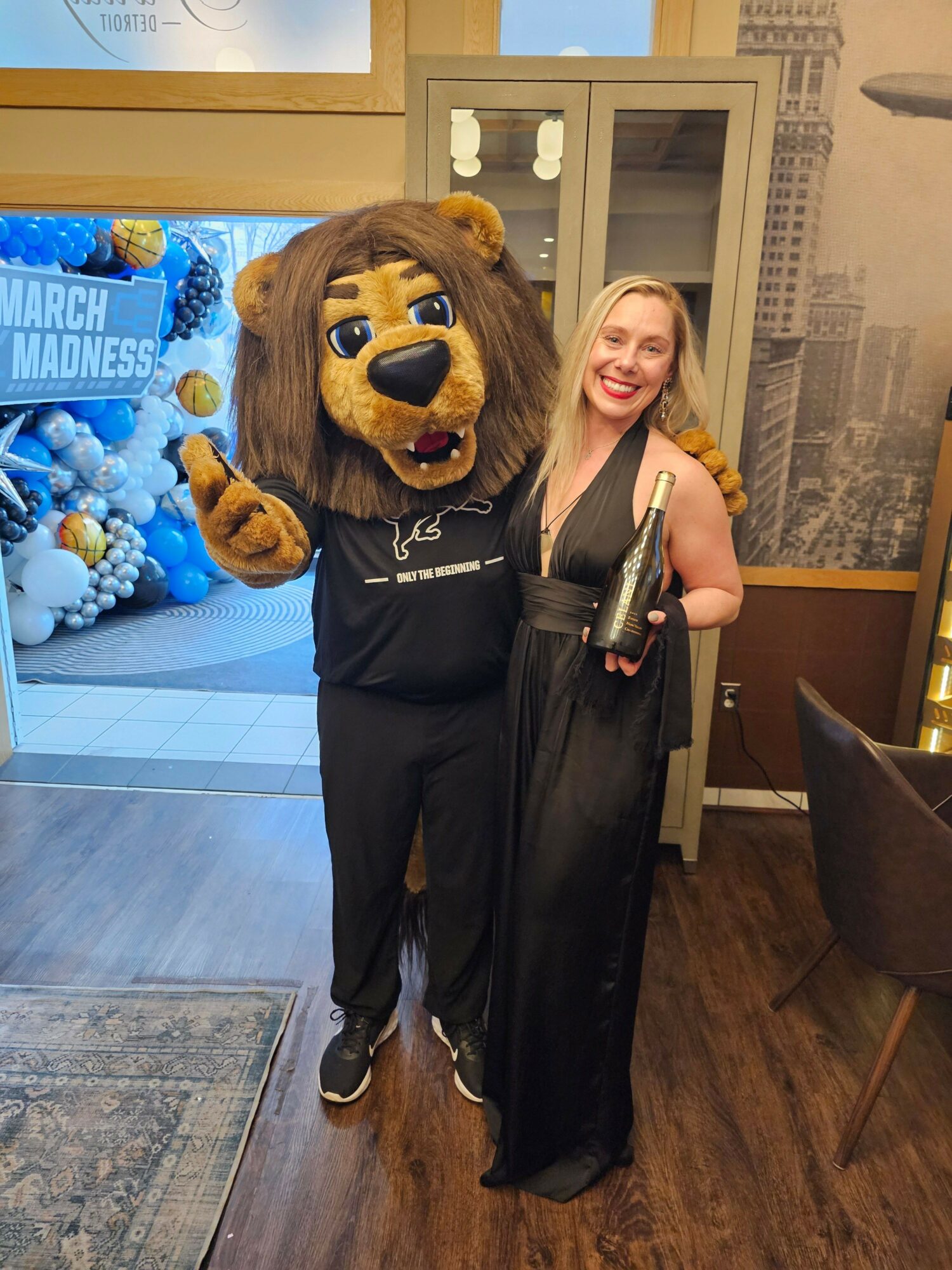 Person in a lion costume standing indoors, holding a microphone, with a woman in a black dress in the background.