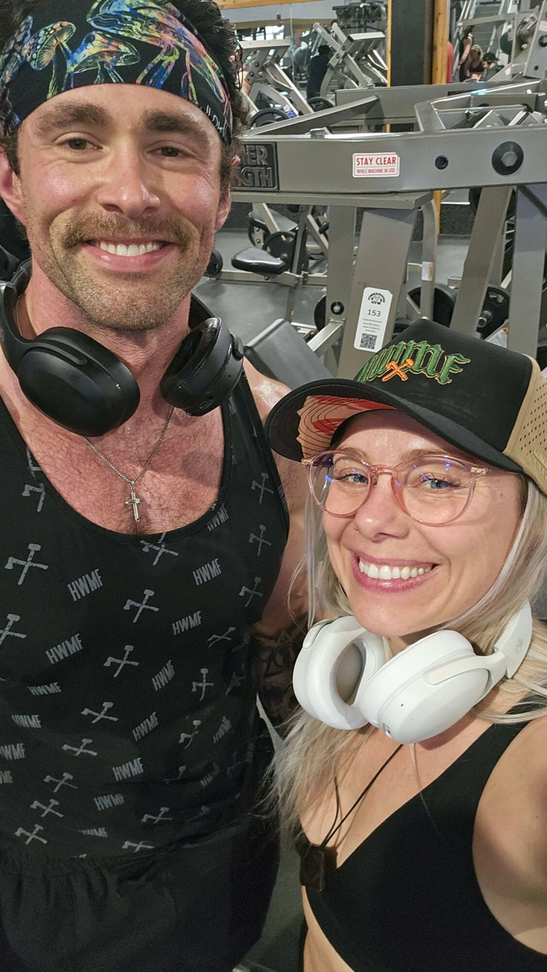 Two smiling people in a gym, one wearing headphones and glasses, the other wearing a cap and a bandana, with gym equipment in the background.