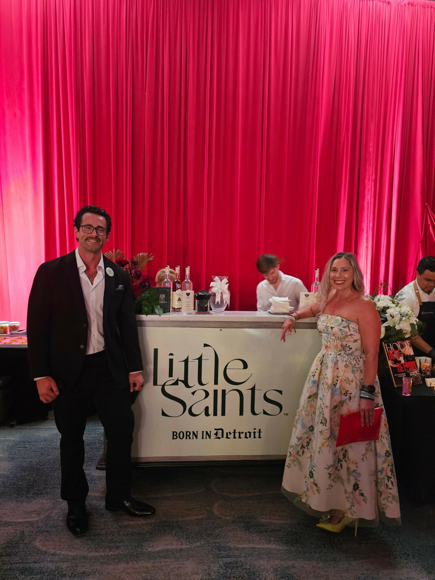 Two people standing in front of a red curtain, one woman in a floral dress and a man in a suit, with a sign reading 'Little Saints'.