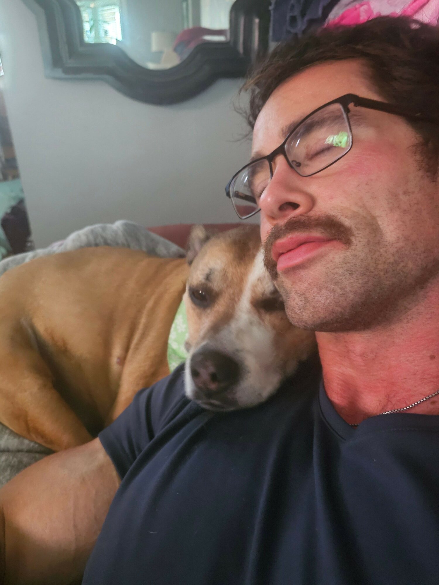 Man with glasses and a dog resting on his shoulder, both lying down, indoors.