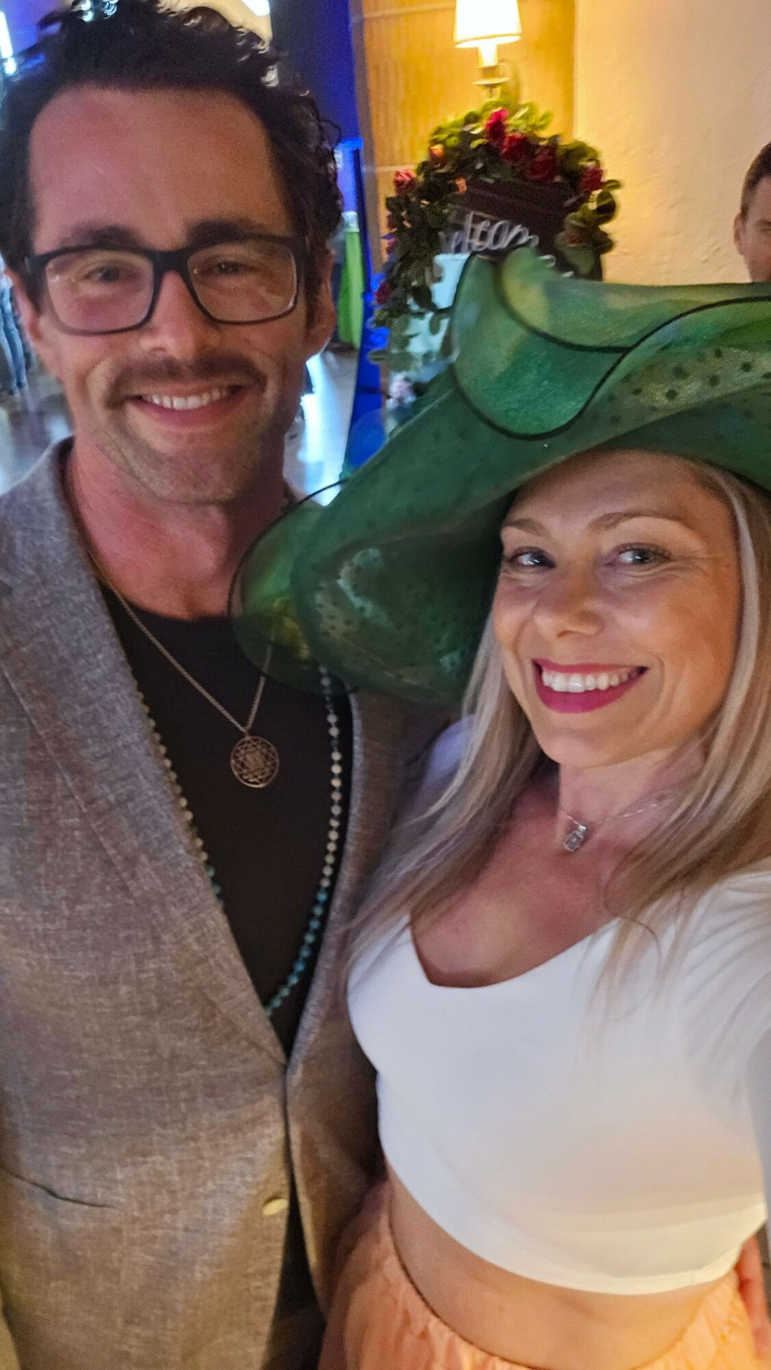 Two smiling people, one wearing glasses and a blazer, the other with long blonde hair and a white top, in a festive setting.