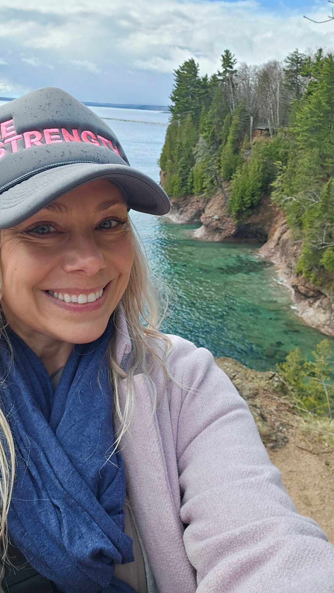 Woman outdoors near a body of water with trees and rocks, smiling, wearing a cap and jacket.