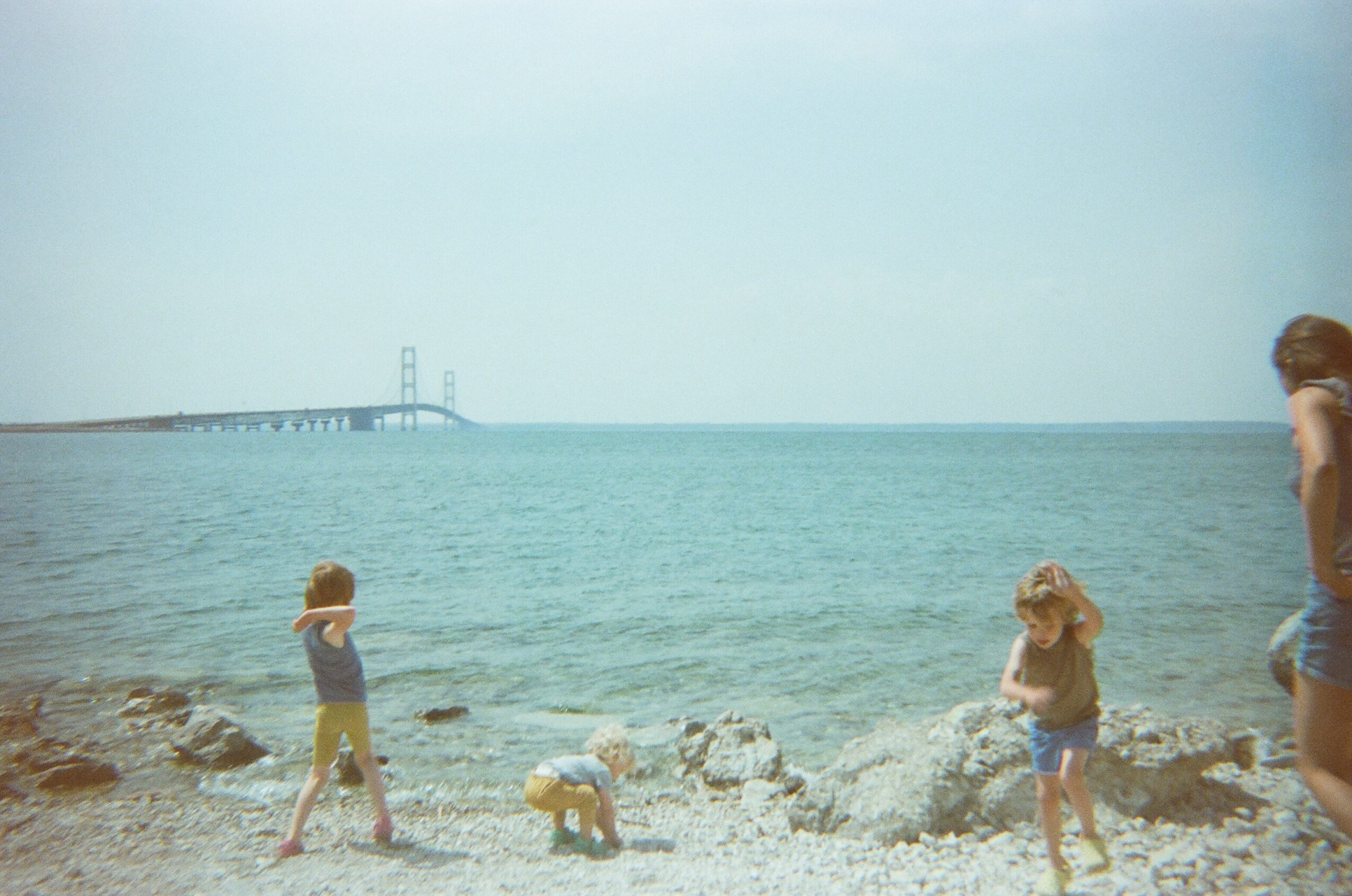Children and a woman on a rocky beach with water and a bridge in the background, clear sky, and calm sea.