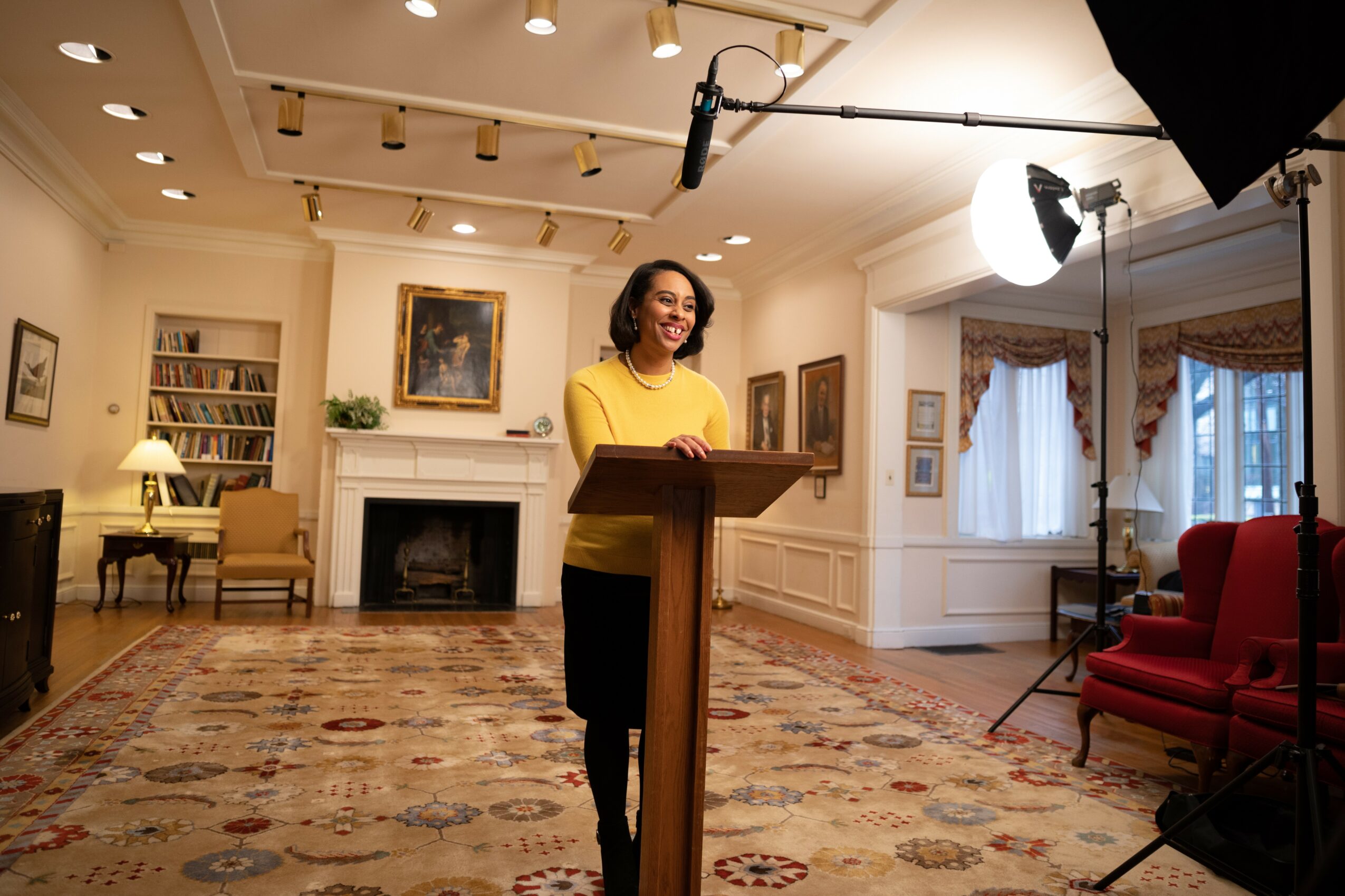 Woman standing at a podium in a spacious, well-lit room with elegant furniture and artwork, preparing to speak.