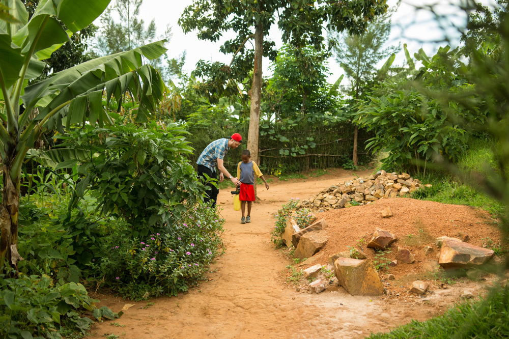 Adult and child walking on a dirt path surrounded by green plants and trees in a garden or park.
