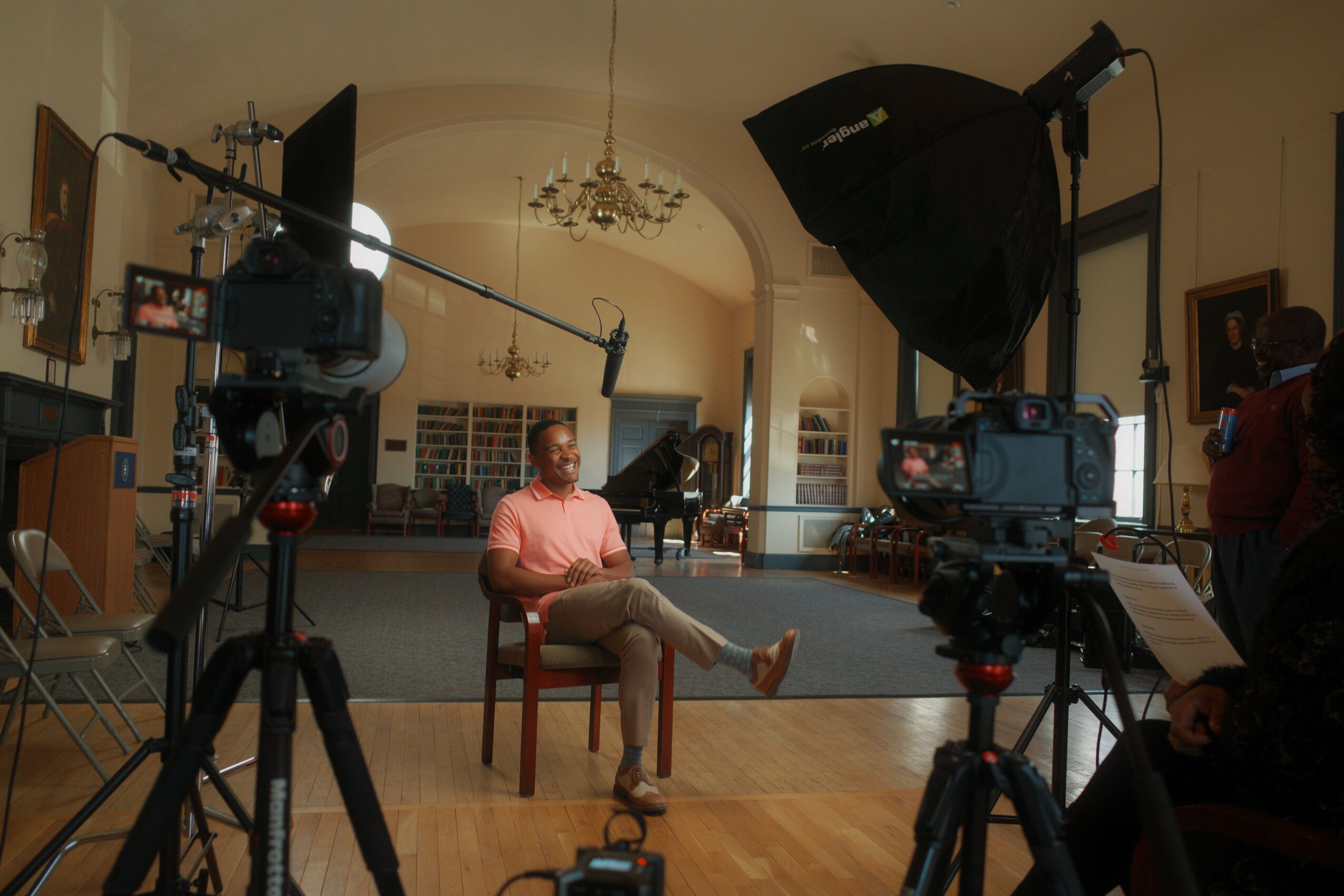 Person sitting on a chair in a room with professional filming equipment, including cameras and lights, in a spacious room with high ceilings.