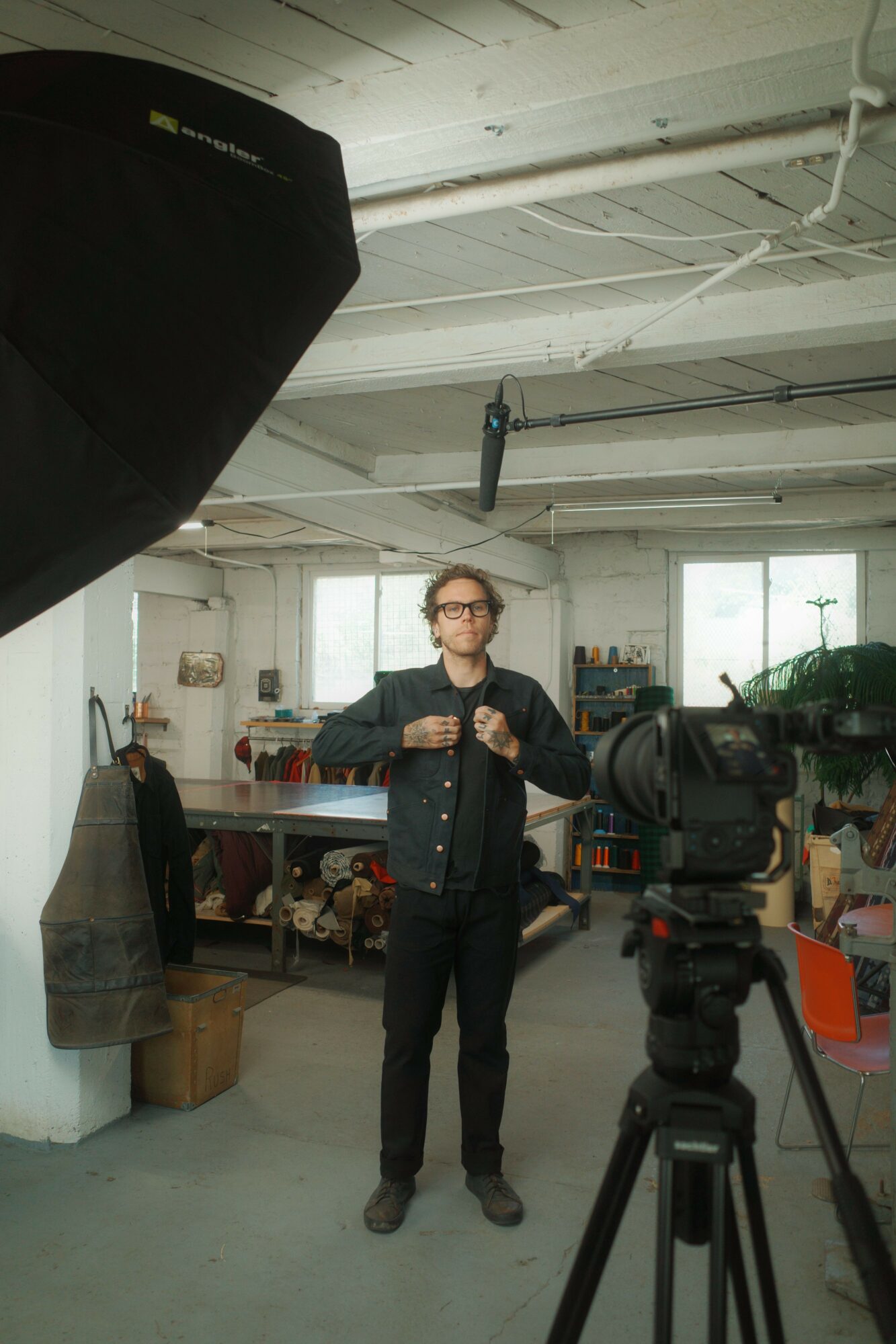 Person standing in a room with camera equipment, shelves, and a window, holding a microphone, facing camera, with a large black object nearby.