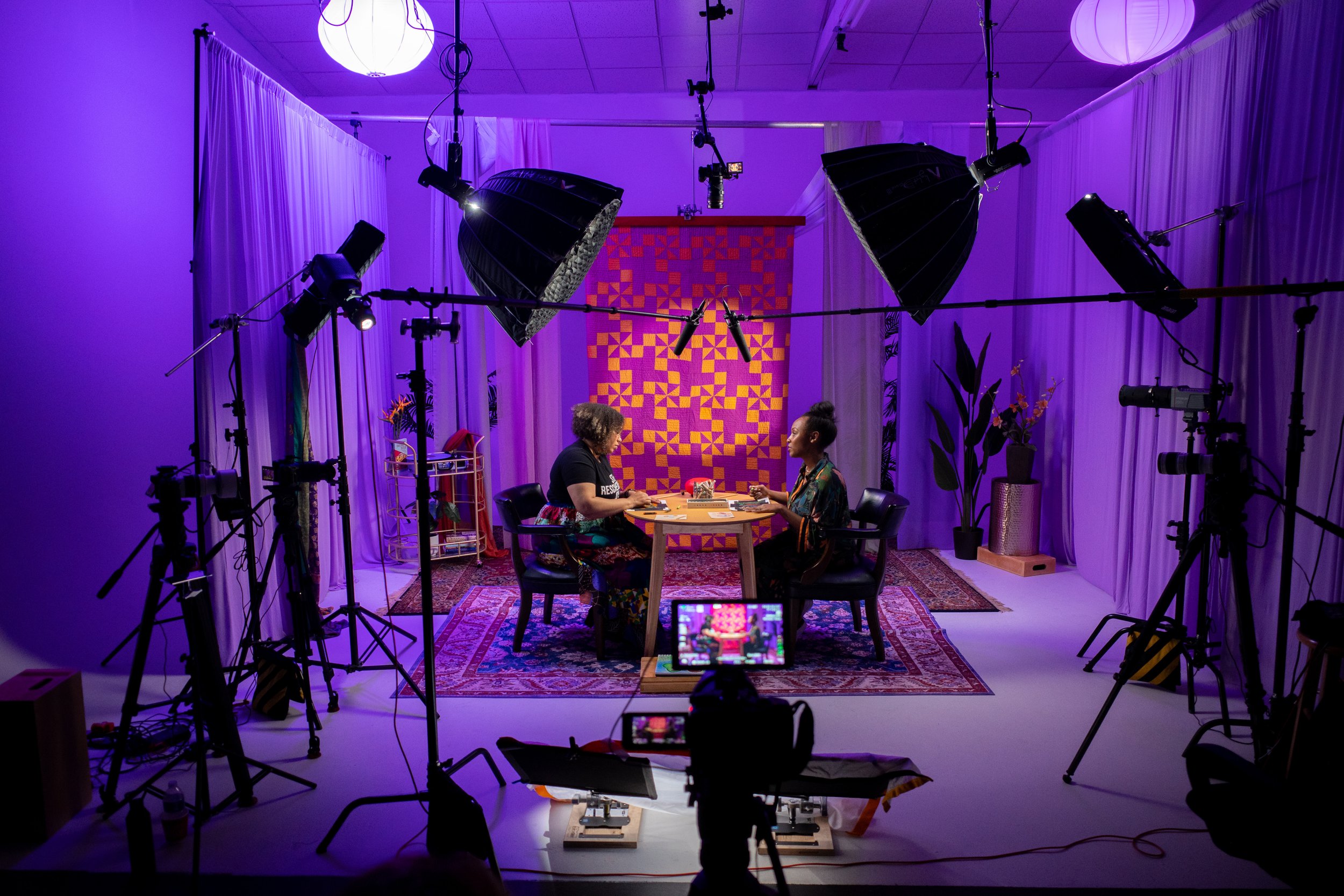 Two people sit at a table in a studio with purple lighting, surrounded by cameras and lighting equipment, and a colorful backdrop.