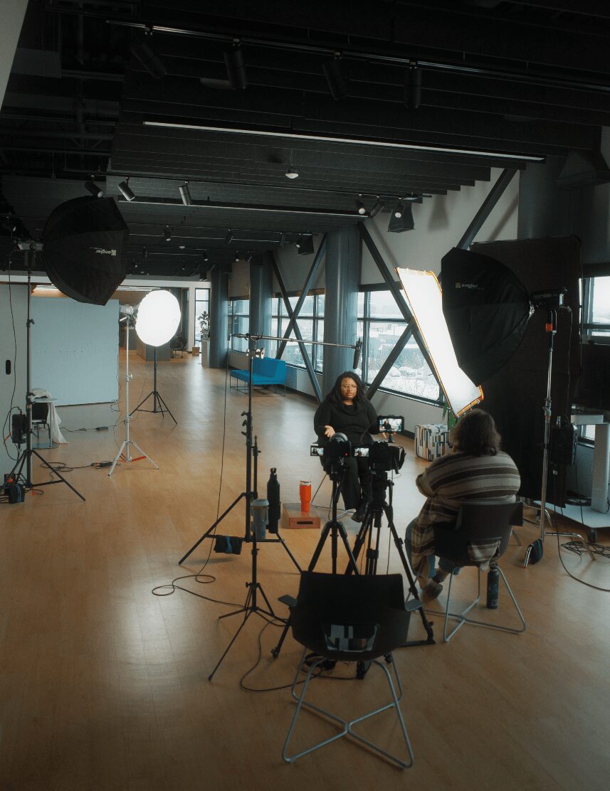 Two people sit in a studio with professional lighting and equipment, engaged in conversation or interview, with a large window in the background.