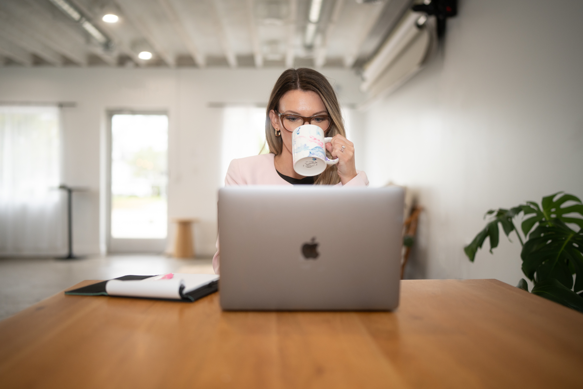 Person with glasses drinking from a mug while working on a laptop at a wooden table, with a plant nearby.