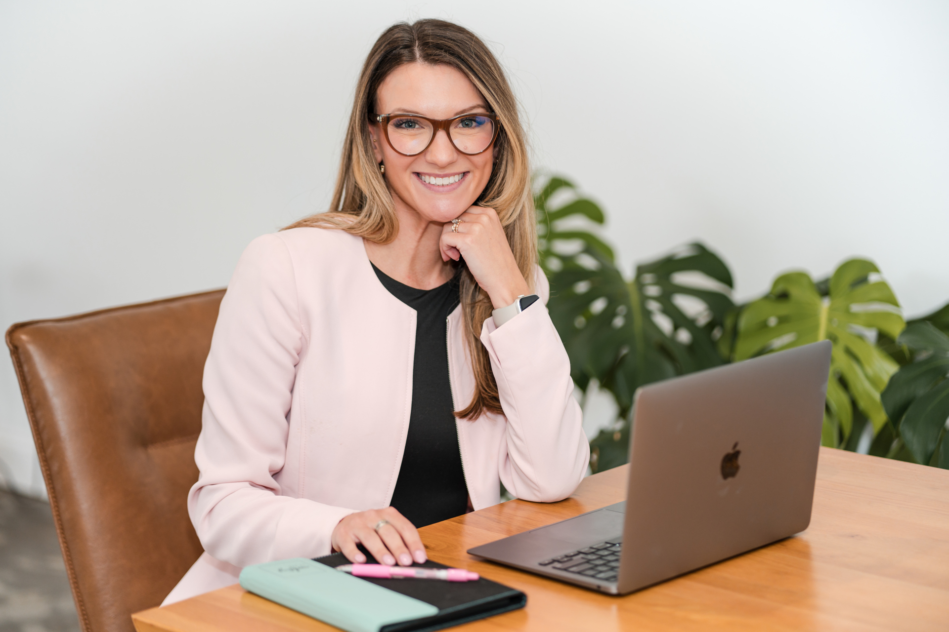 Woman with glasses smiling, sitting at a desk with a laptop and a notebook, in front of green plants.