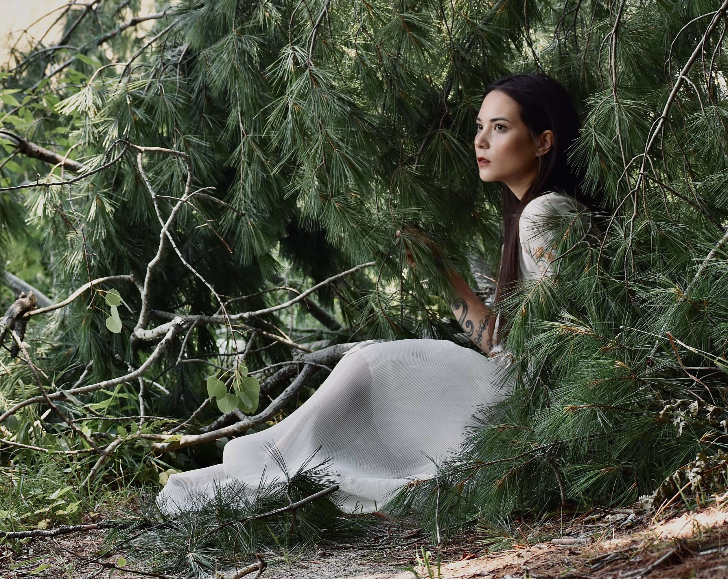 Woman in white dress sitting among green tree branches and foliage, looking to the side.