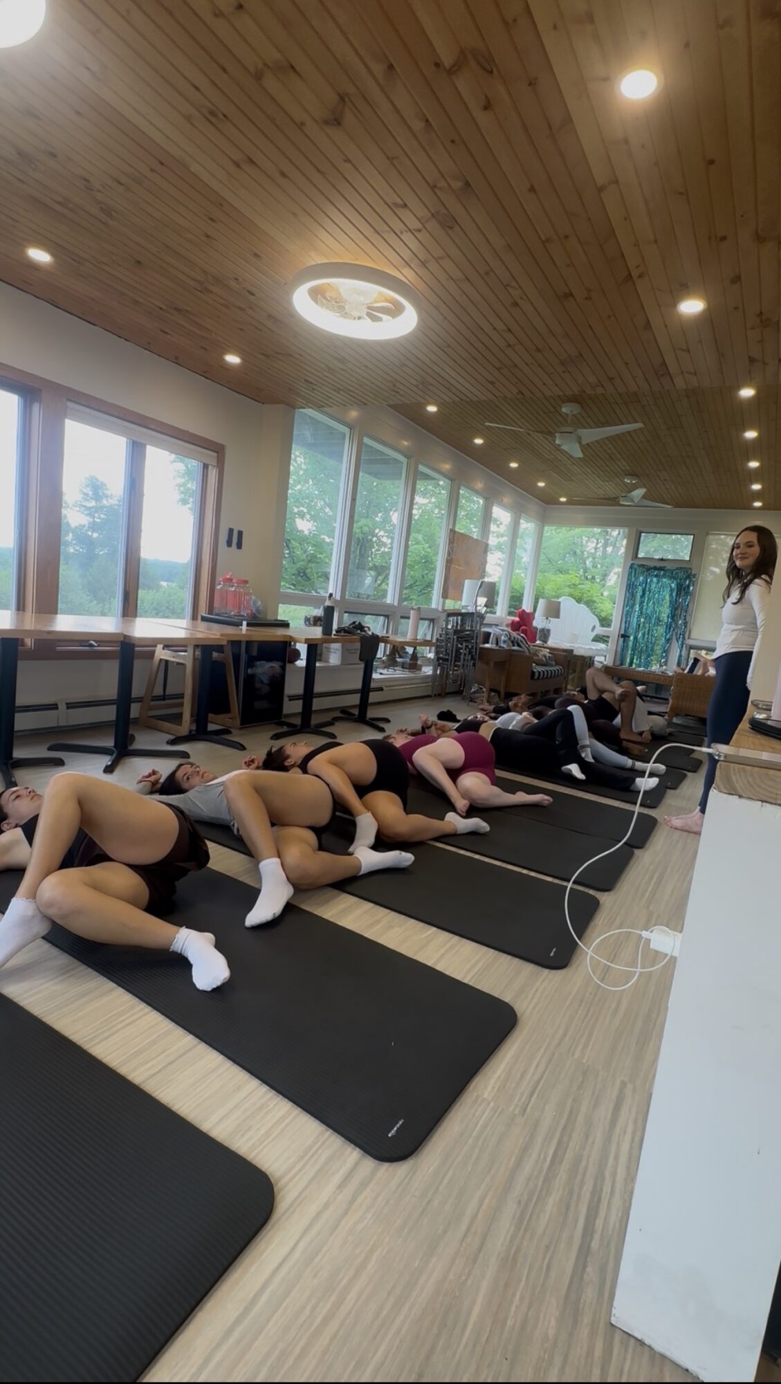 Group of people lying on mats in a room with large windows and wooden ceiling, instructor standing nearby, bright natural light.