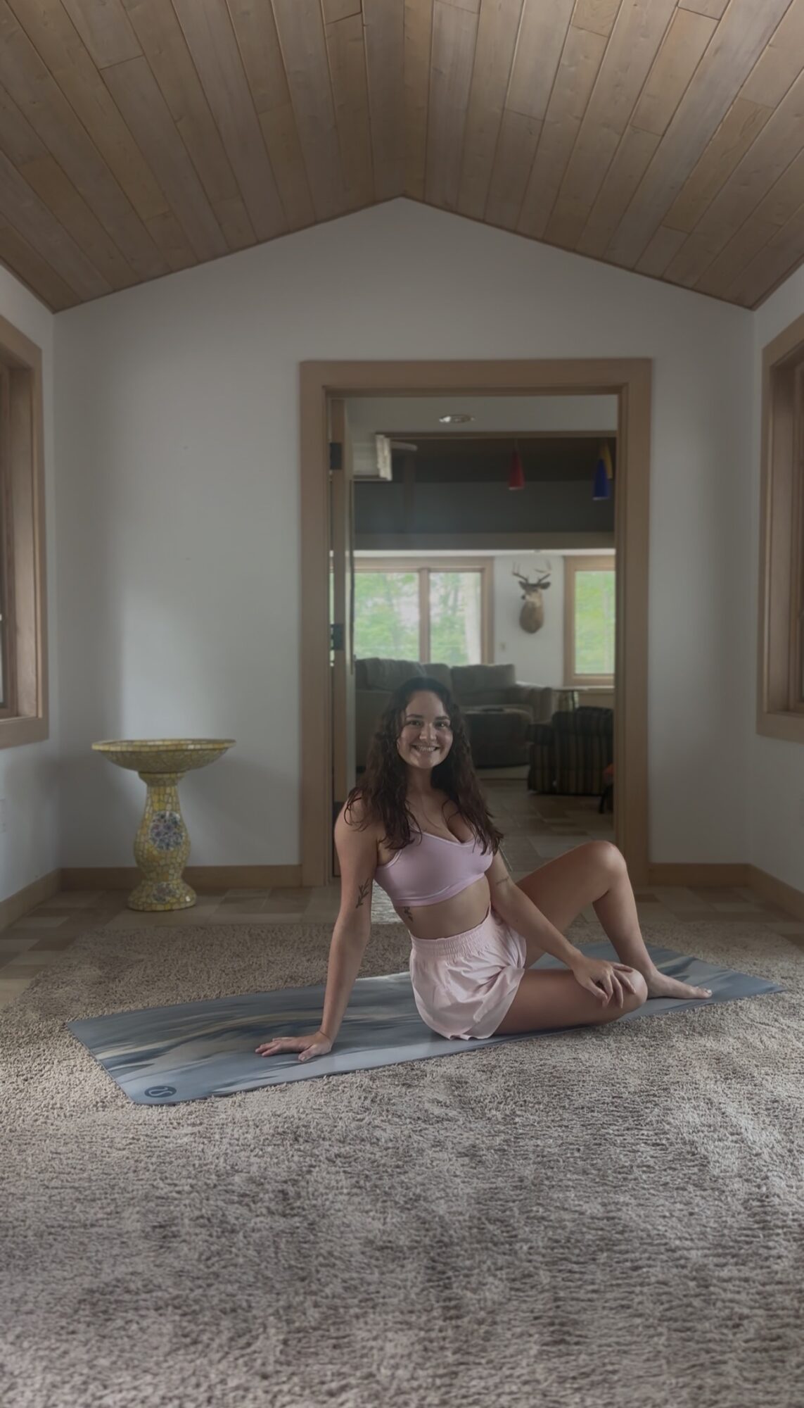 Young woman sitting on a yoga mat in a room with wooden ceiling, white walls, and large windows, smiling.