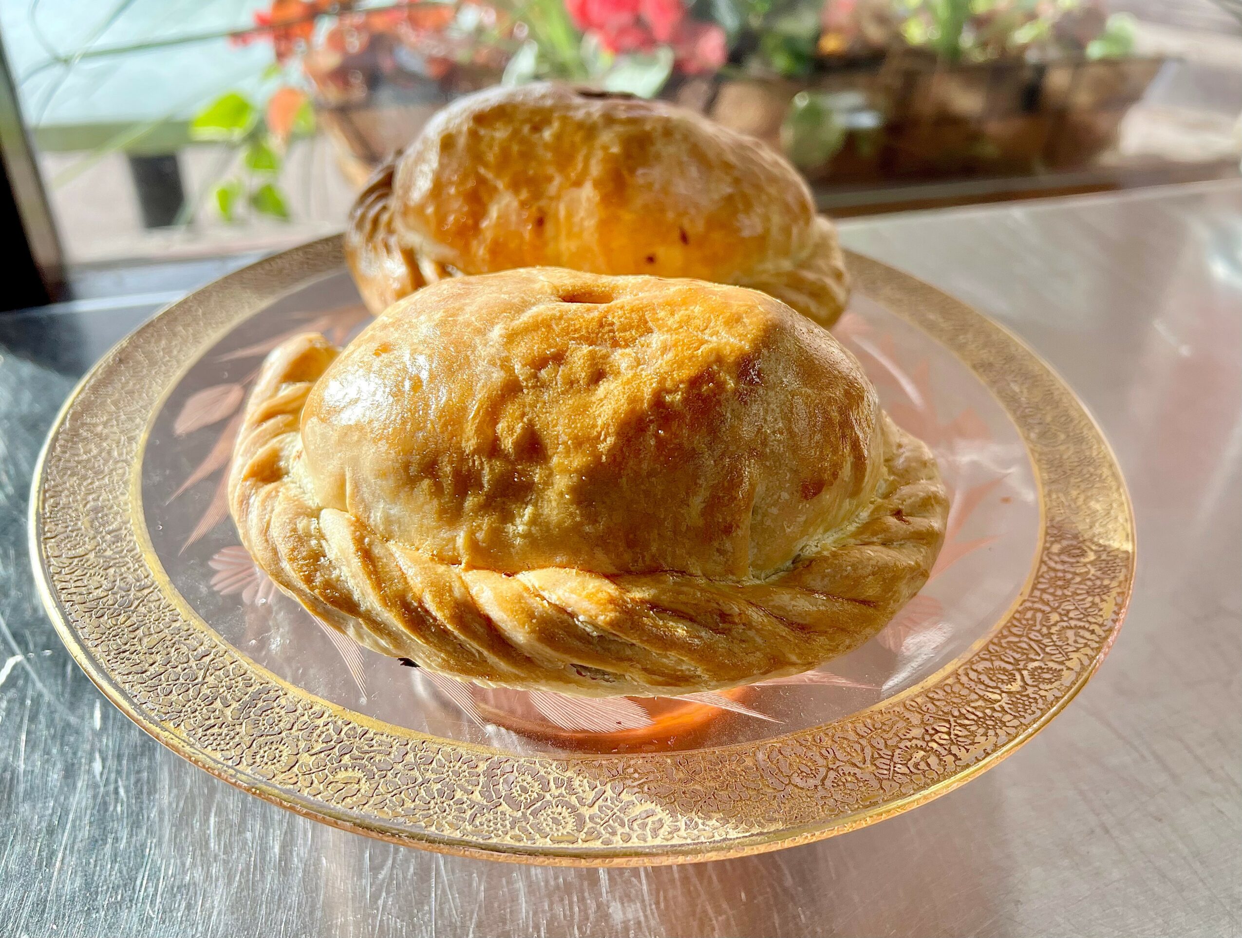 Two baked pastries on a decorative glass plate with a floral background, sunlight shining on them.
