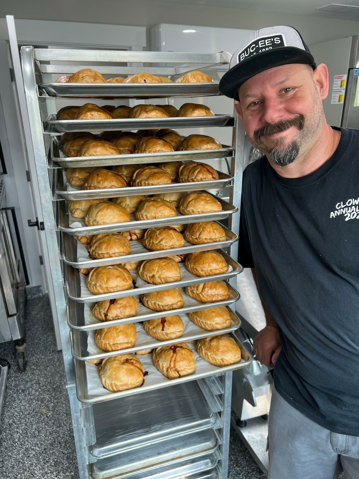 Man with a beard and black cap stands next to a tray of baked goods on a metal rack.