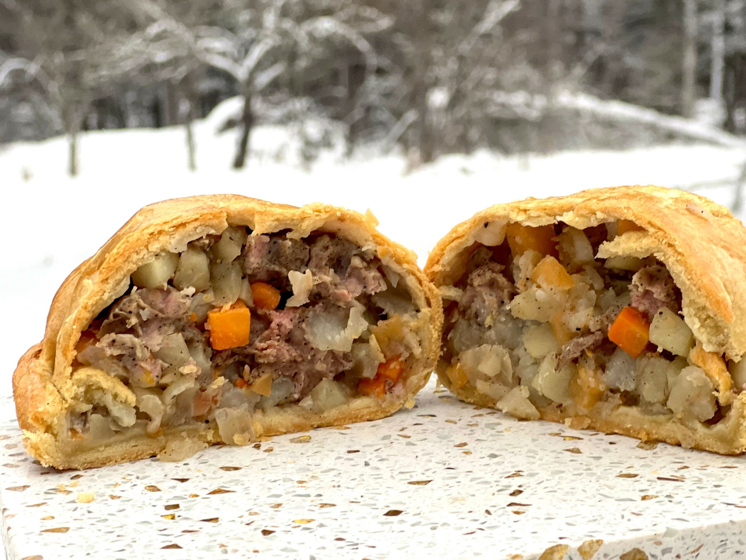 Cross-section of a meat and vegetable-filled pastry with visible carrots, potatoes, and meat, outdoors with snow-covered background.