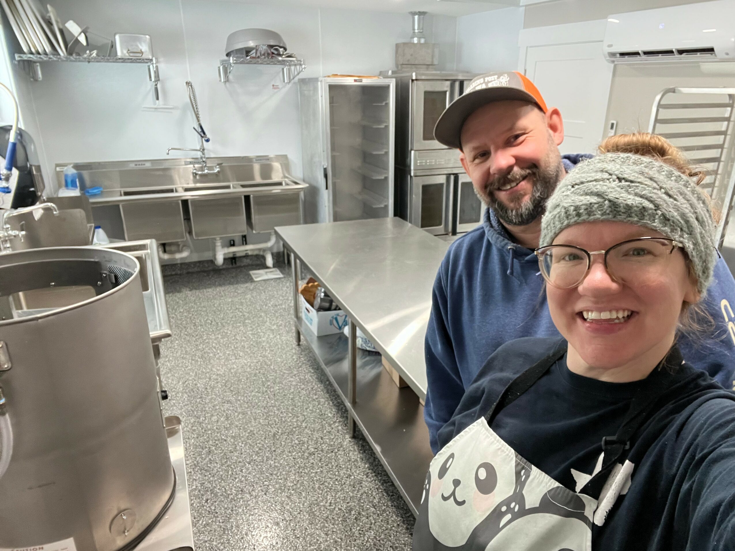 Two people smiling in a commercial kitchen, one wearing a cap and the other a headband, with stainless steel appliances around them.