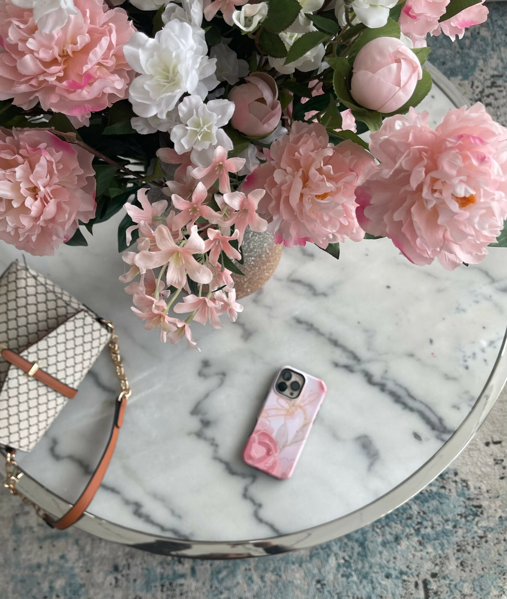 Vase with pink and white flowers on marble table, phone with pink floral case, and patterned bag nearby.