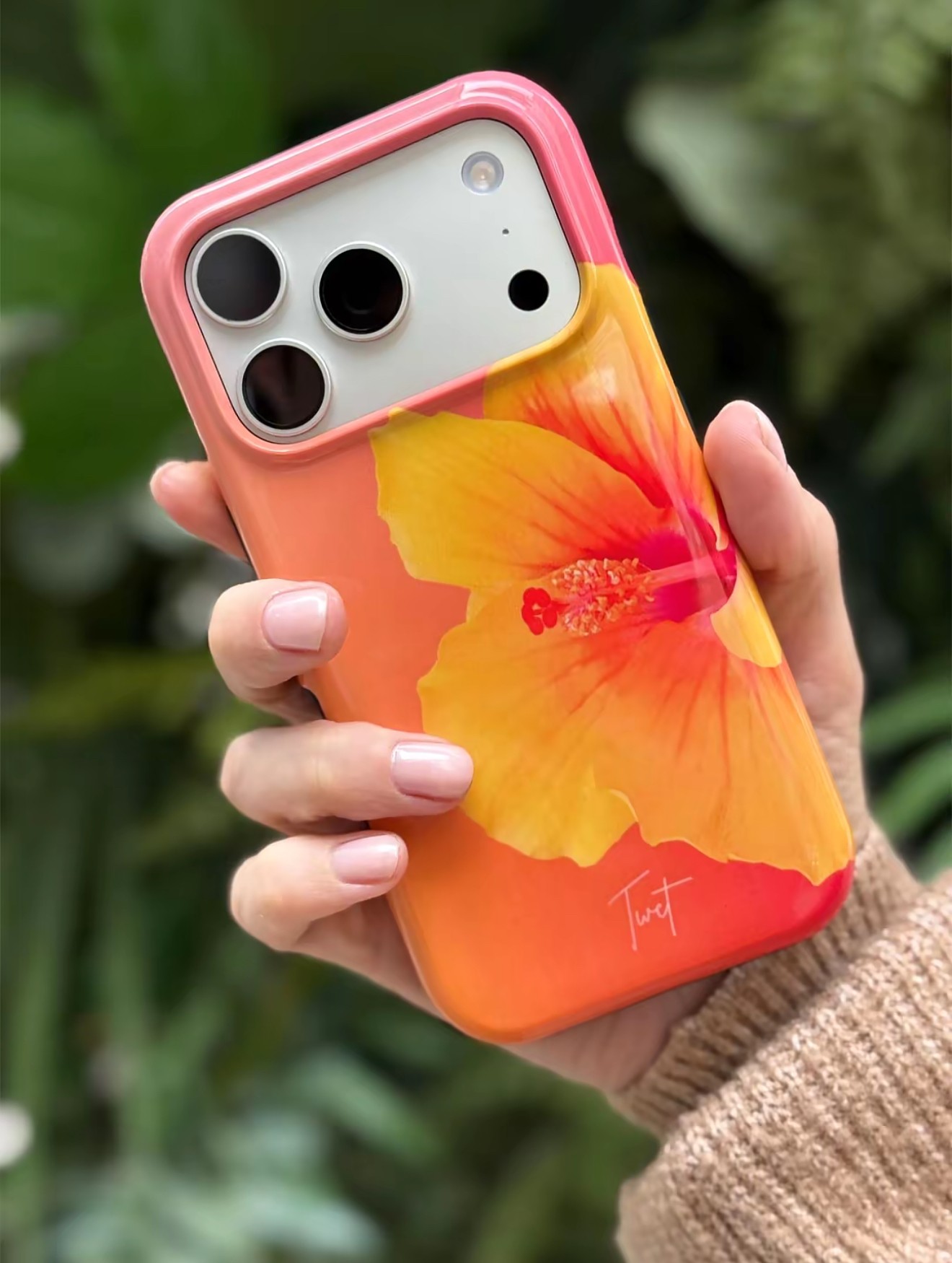 Hand holding a smartphone with a colorful flower design case, background of green foliage, person wearing a beige sweater