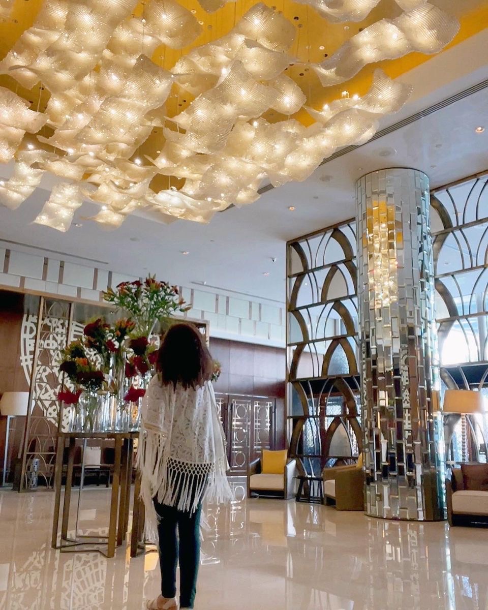 Person with long hair and fringed shawl stands in a decorated room with floral arrangements and modern furniture.