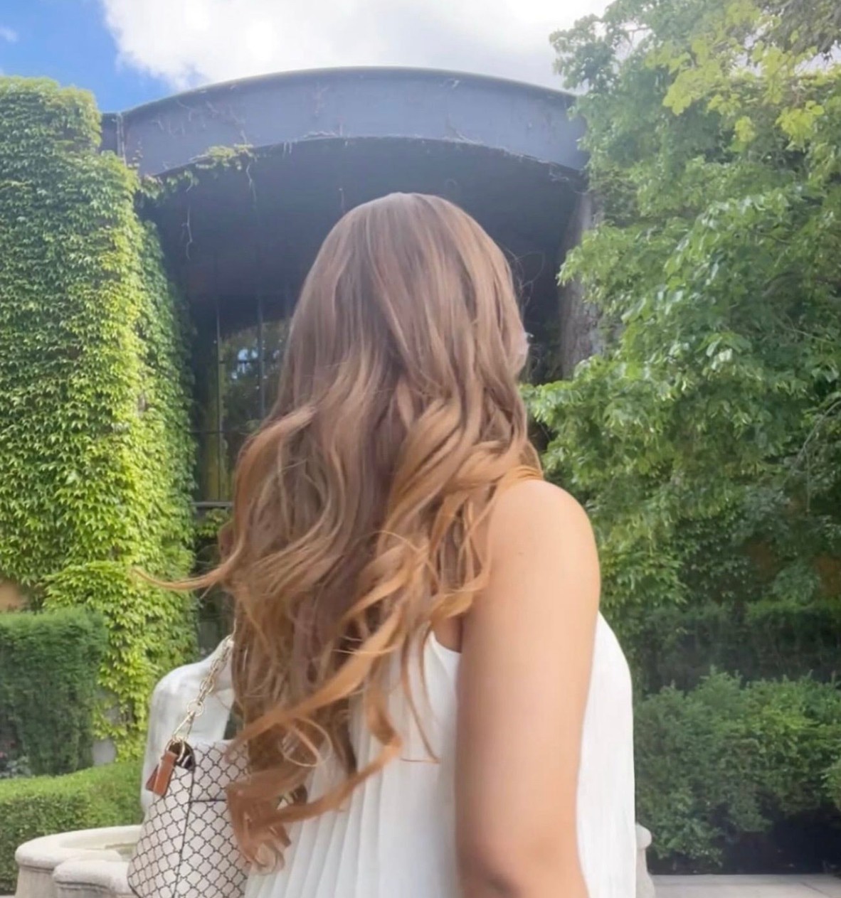 Back view of a woman with long, wavy, light brown hair outdoors, with greenery and a bridge in the background.
