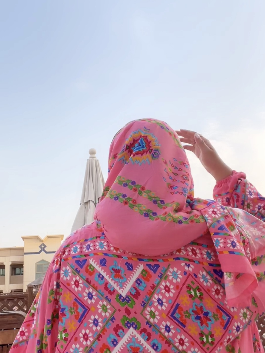 Person wearing a colorful, patterned headscarf, facing away, with a building and umbrella in the background.
