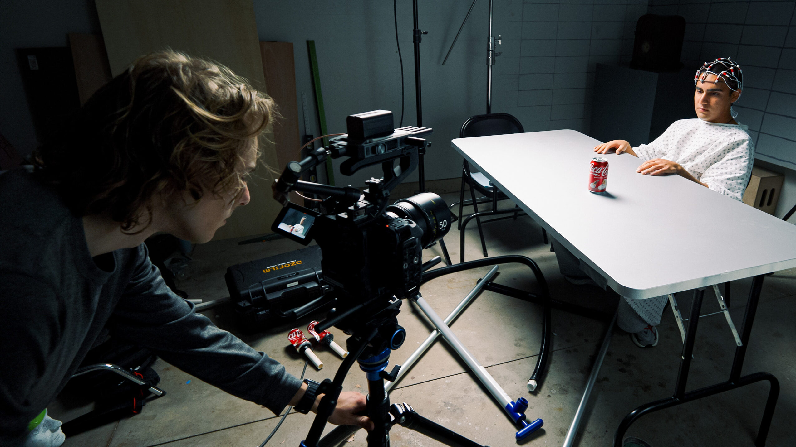 Person filming a child sitting at a table with a red cup in a room with a white wall and equipment.